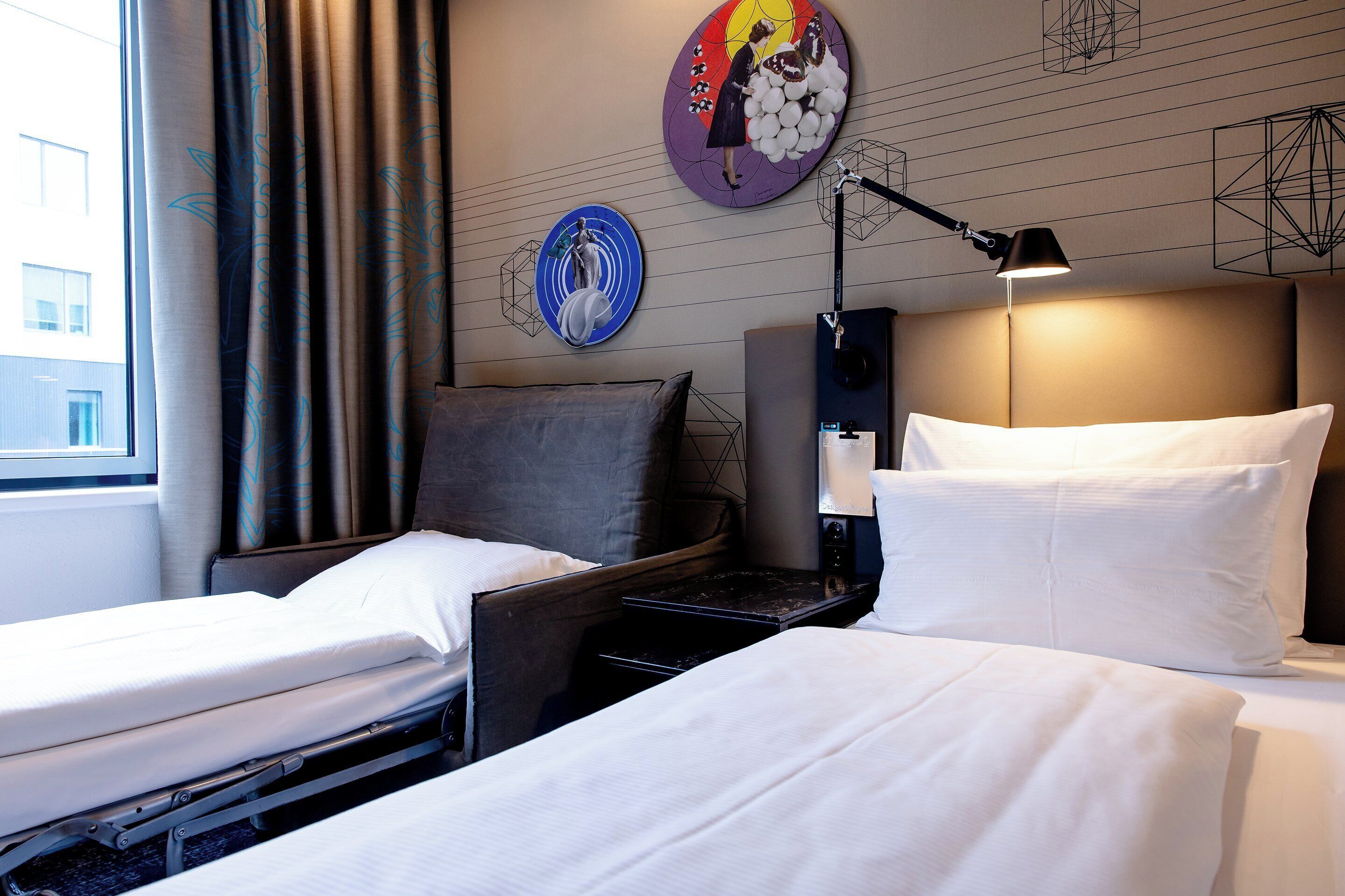 Ein Hotelzimmer mit zwei Einzelbetten, einem Nachttisch, einer Lampe und einer Wand mit Kunstwerken.