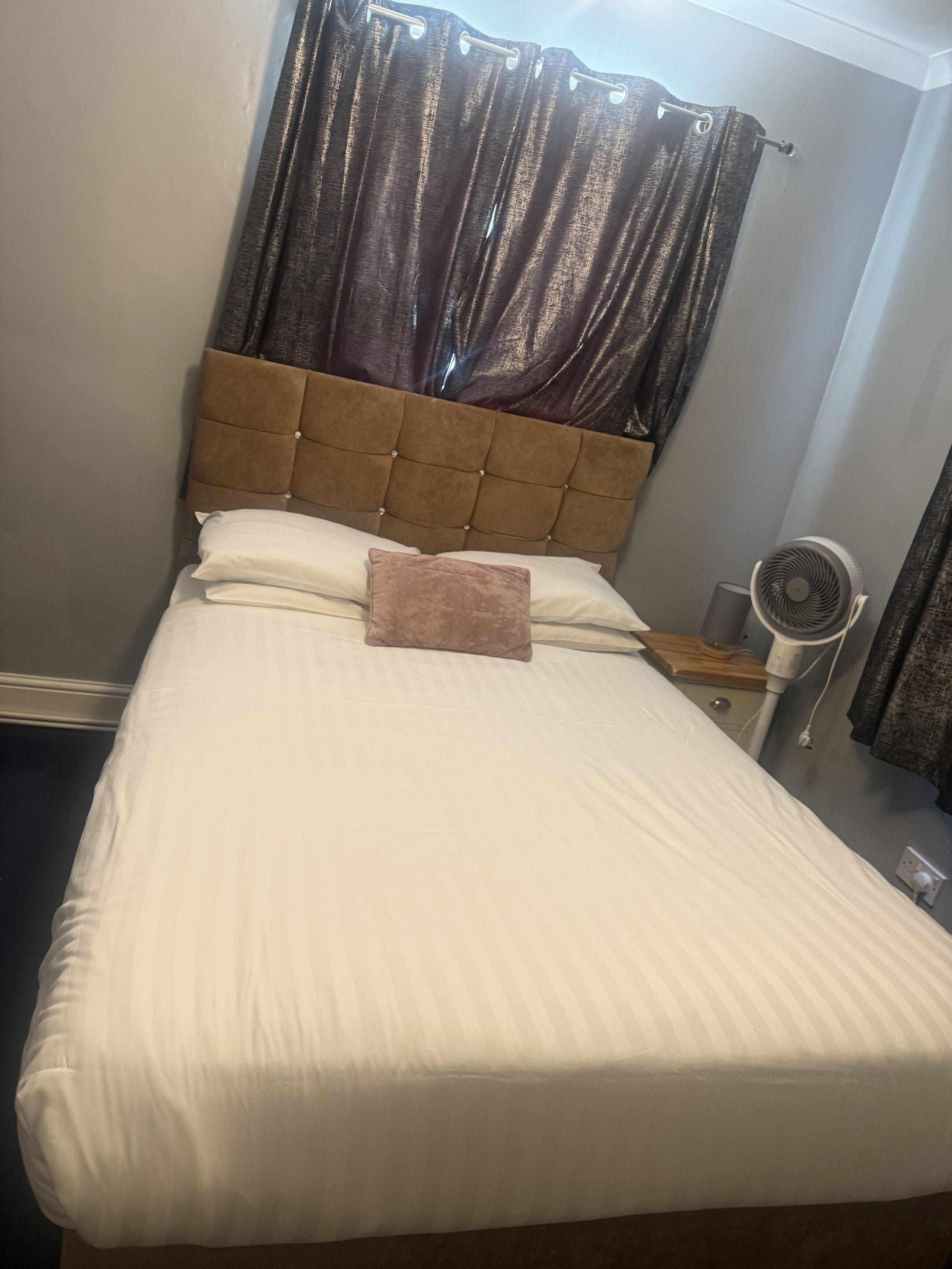 A neatly made bed with white linens and a brown headboard, a bedside table with a fan, and a window with dark curtains.