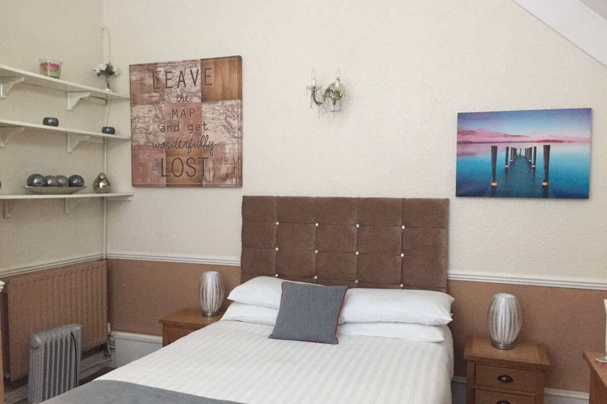 Une chambre à coucher avec un grand lit, une tête de lit, des tables de chevet, une étagère avec des objets décoratifs et un mur orné d’une citation inspirante et d’un tableau.