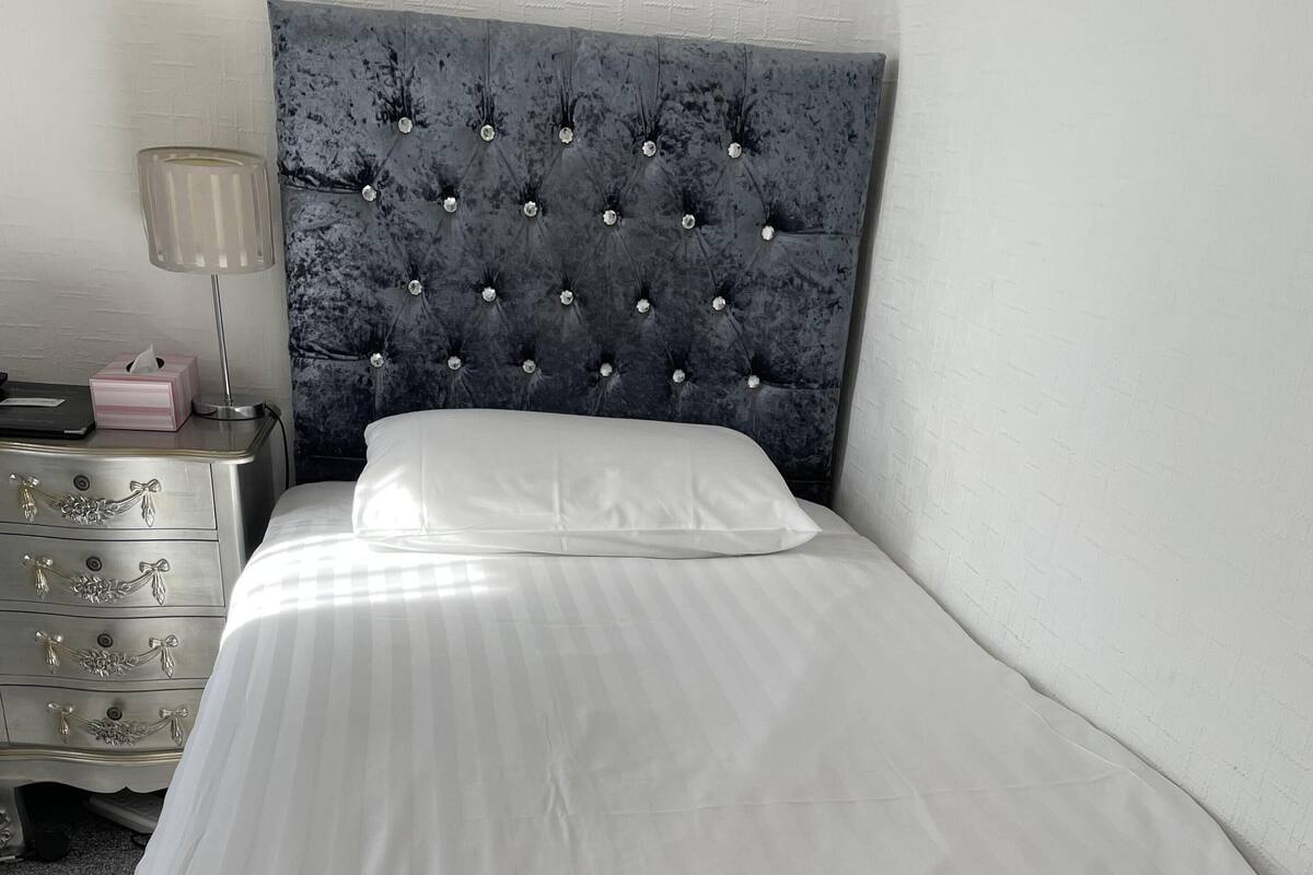 Un lit bien fait, avec une serviette pliée, une tête de lit capitonnée et une table de chevet sur laquelle se trouvent une lampe et une boîte de mouchoirs.