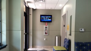 A hotel lobby with a television mounted on the wall, a fire extinguisher, a trash can, and a seating area with a blue and yellow chair. - Cityview Inn & Suites Downtown RiverCenter Area (San Antonio)
