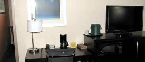Habitación de hotel con escritorio, una silla, televisor, cafetera y un cuadro abstracto enmarcado en la pared.