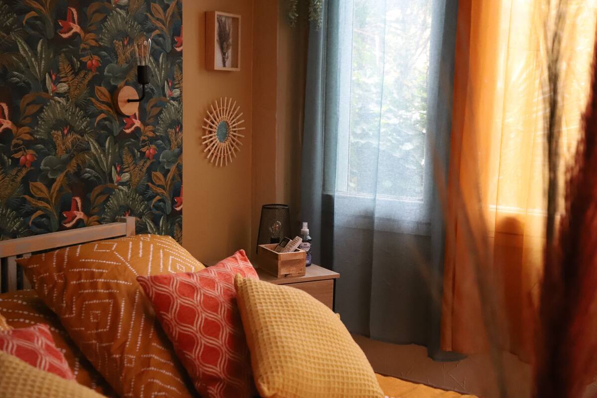 A bedroom with a bed, pillows, a nightstand, a wall clock, a framed picture, and a window with curtains.
