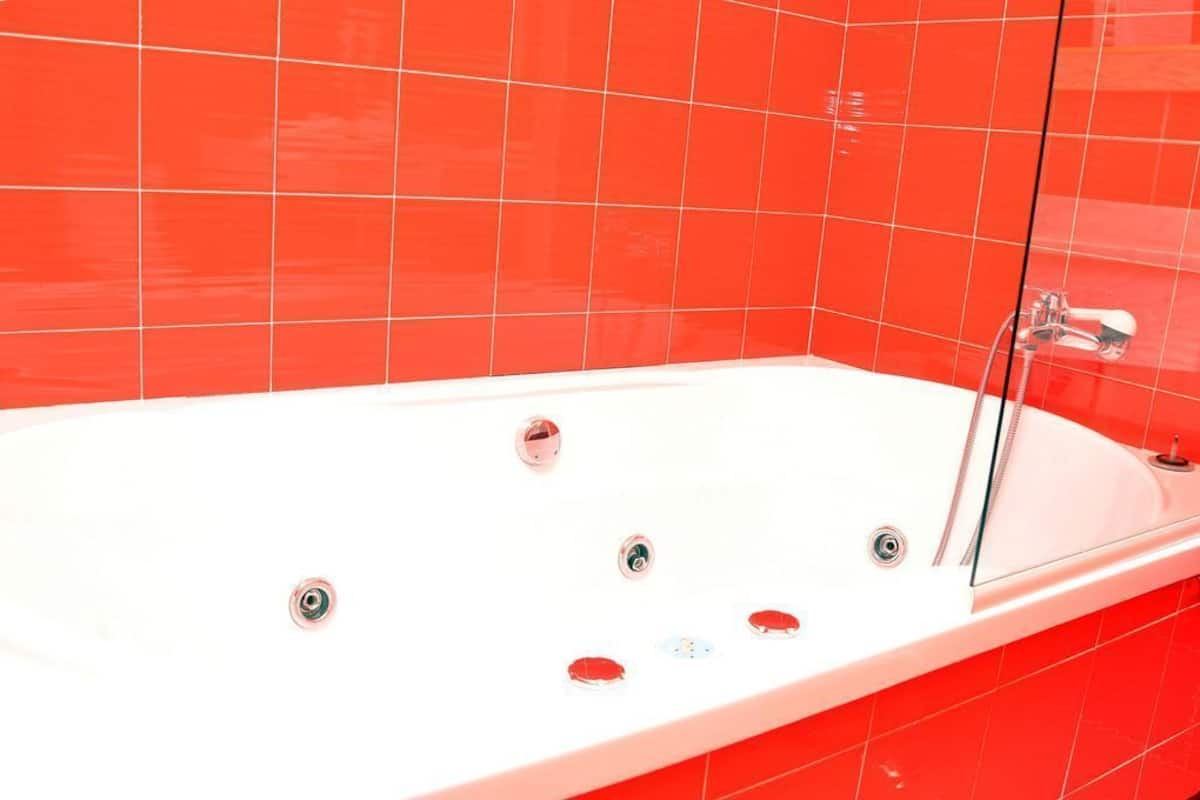 A white bathtub with a glass door, set against a red tiled wall.