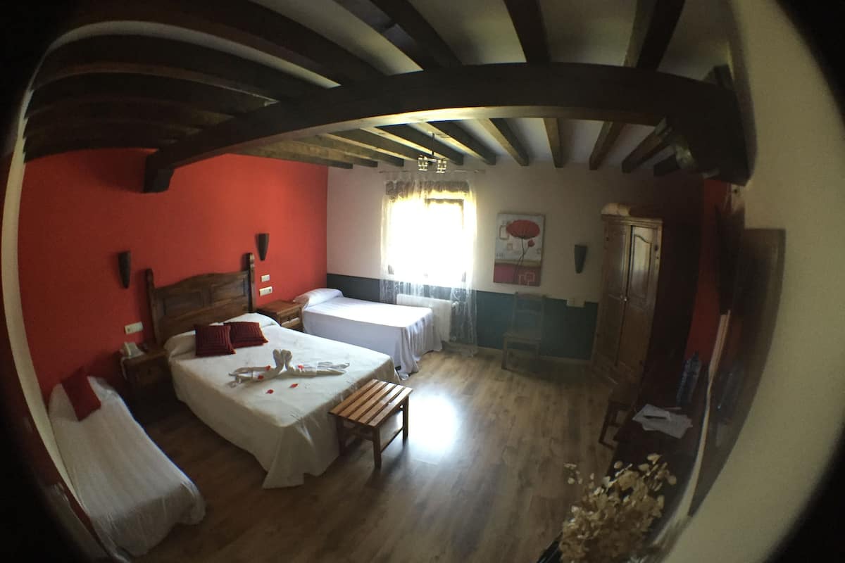 A room with two beds, wooden flooring, a wooden bench, a window with curtains, and a wall with a red and white design.