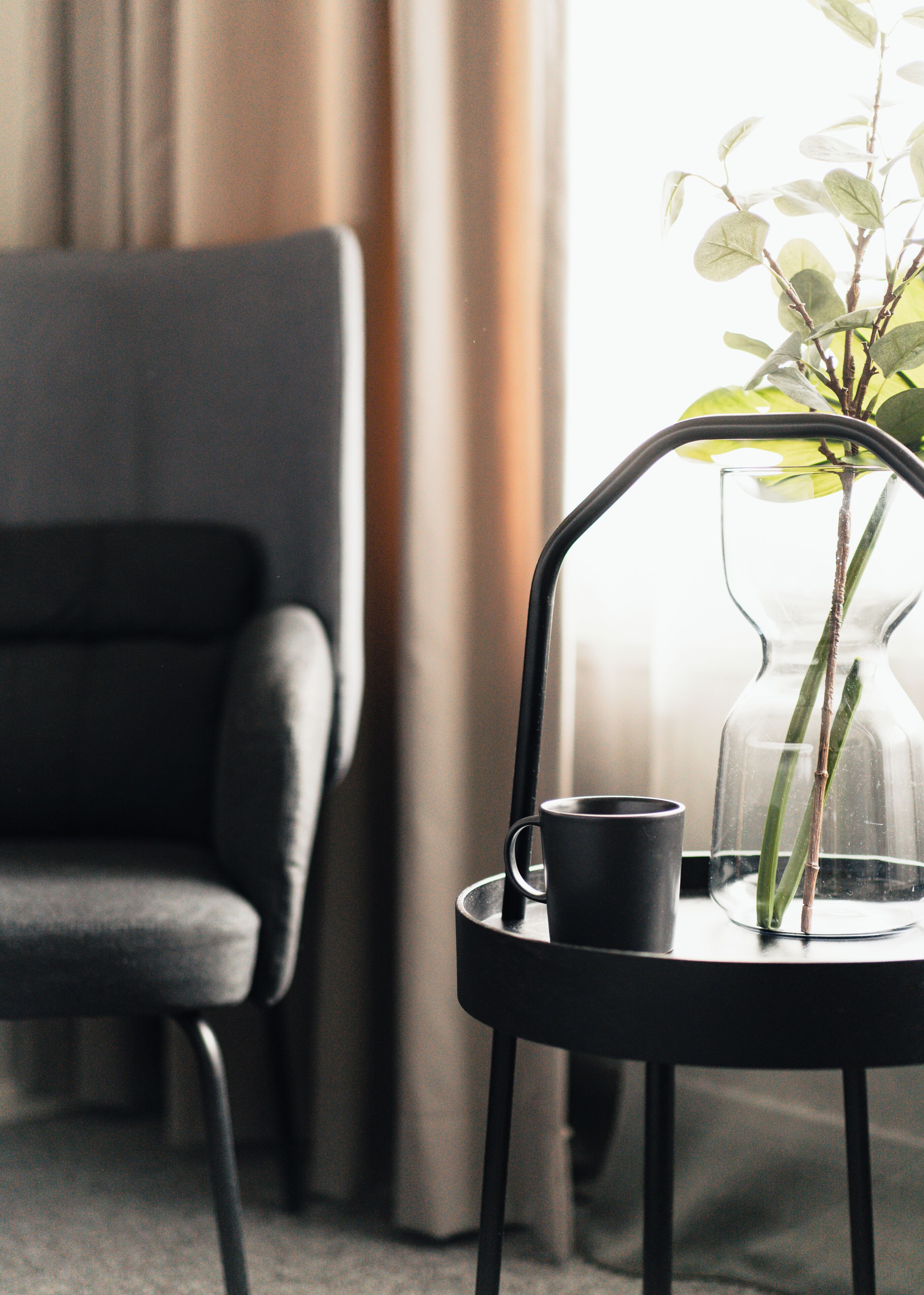 A modern room with a black chair, a clear glass vase with a plant, and a black mug on a small table.