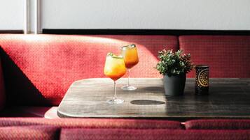 A red upholstered bench with a small table in front. On the table, there are two glasses of orange drink, a small potted plant, and a decorative candle holder.