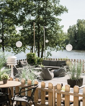 An outdoor seating area with a view of a lake, featuring wooden tables, chairs, and a sofa. - Hotel Waltikka (Valkeakoski)