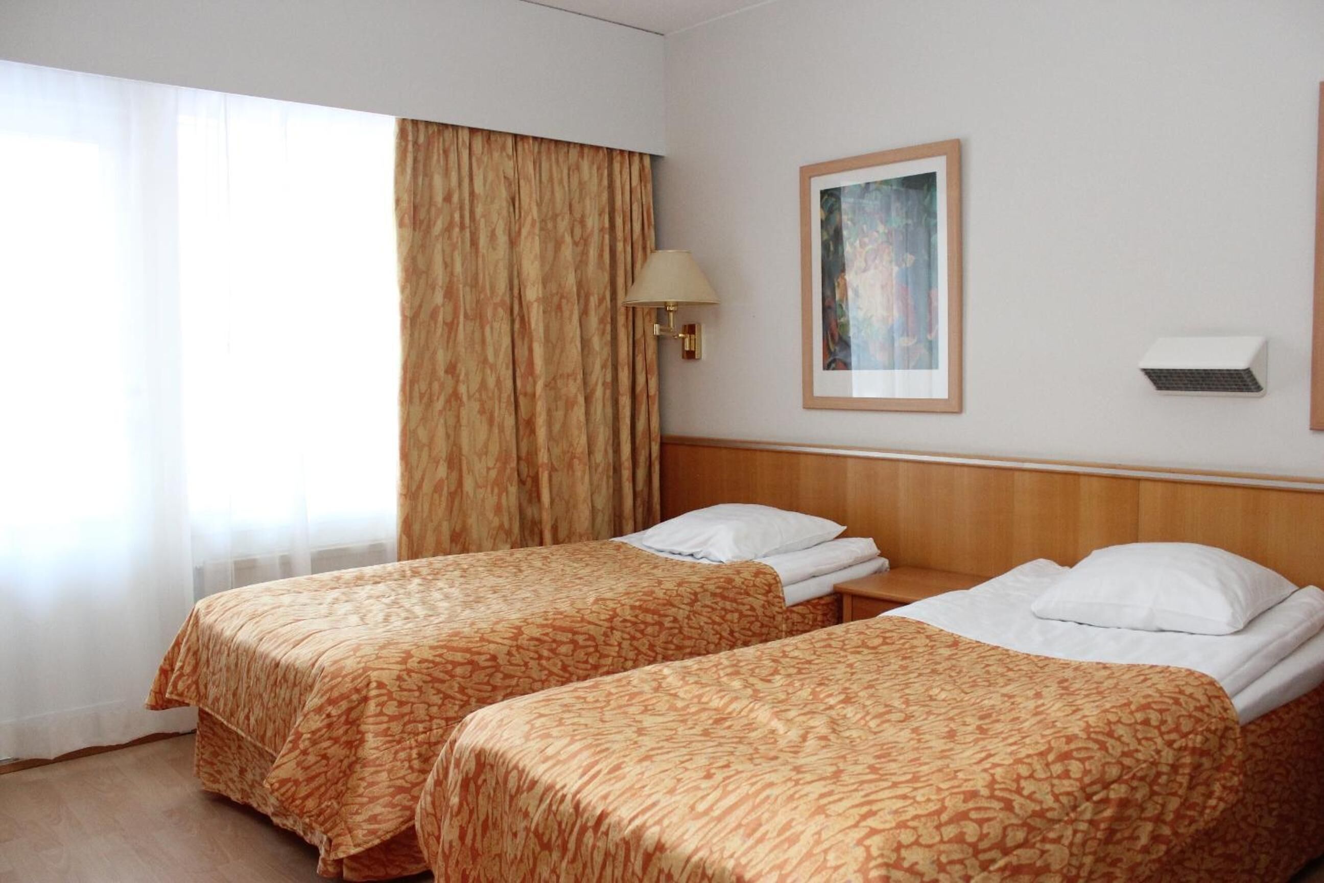 Habitación de hotel con dos camas, una ventana con cortinas translúcidas, una lámpara de pared y un cuadro enmarcado.