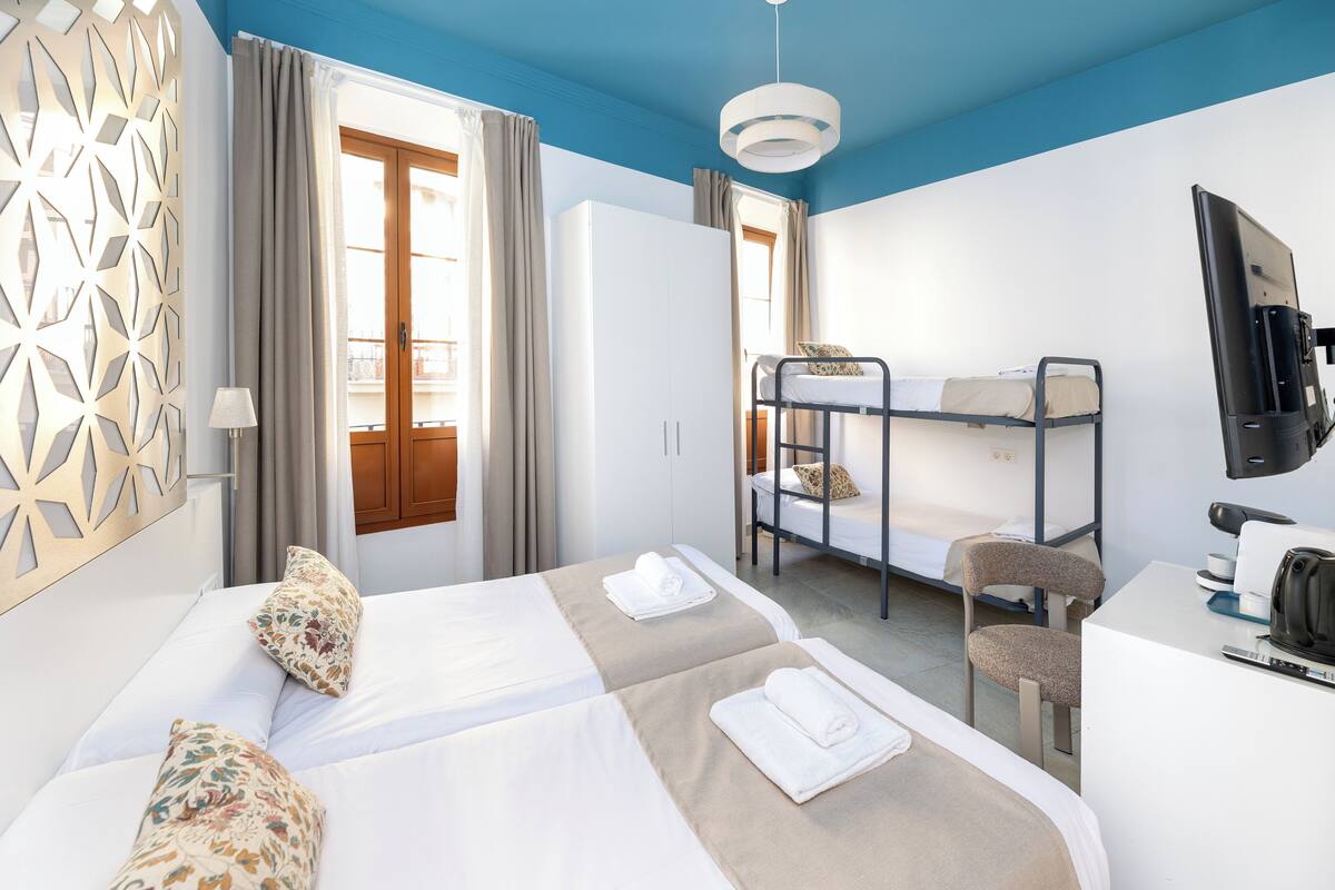 A hotel room with two beds, a bunk bed, a desk with a computer, a TV, and a decorative wall panel.
