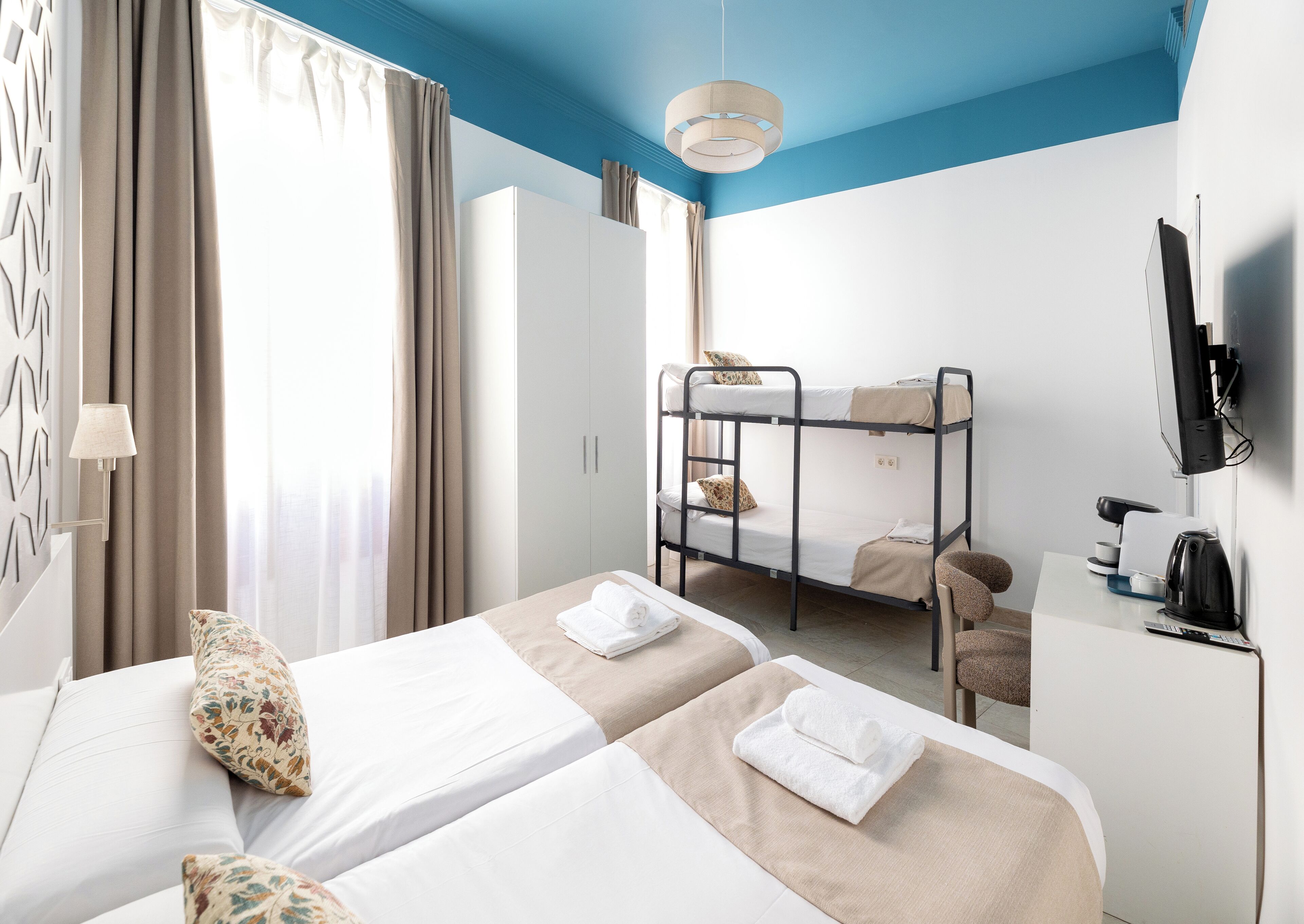 A hotel room with a bunk bed, a desk with a kettle, a TV, and a bed with a pillow.