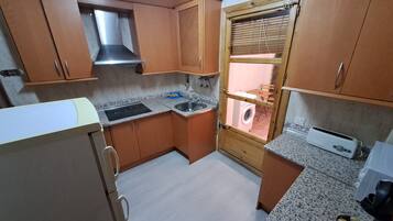Een kleine keuken met houten kastjes, een inbouwoven, een spoelbak en een raam met zonwering.