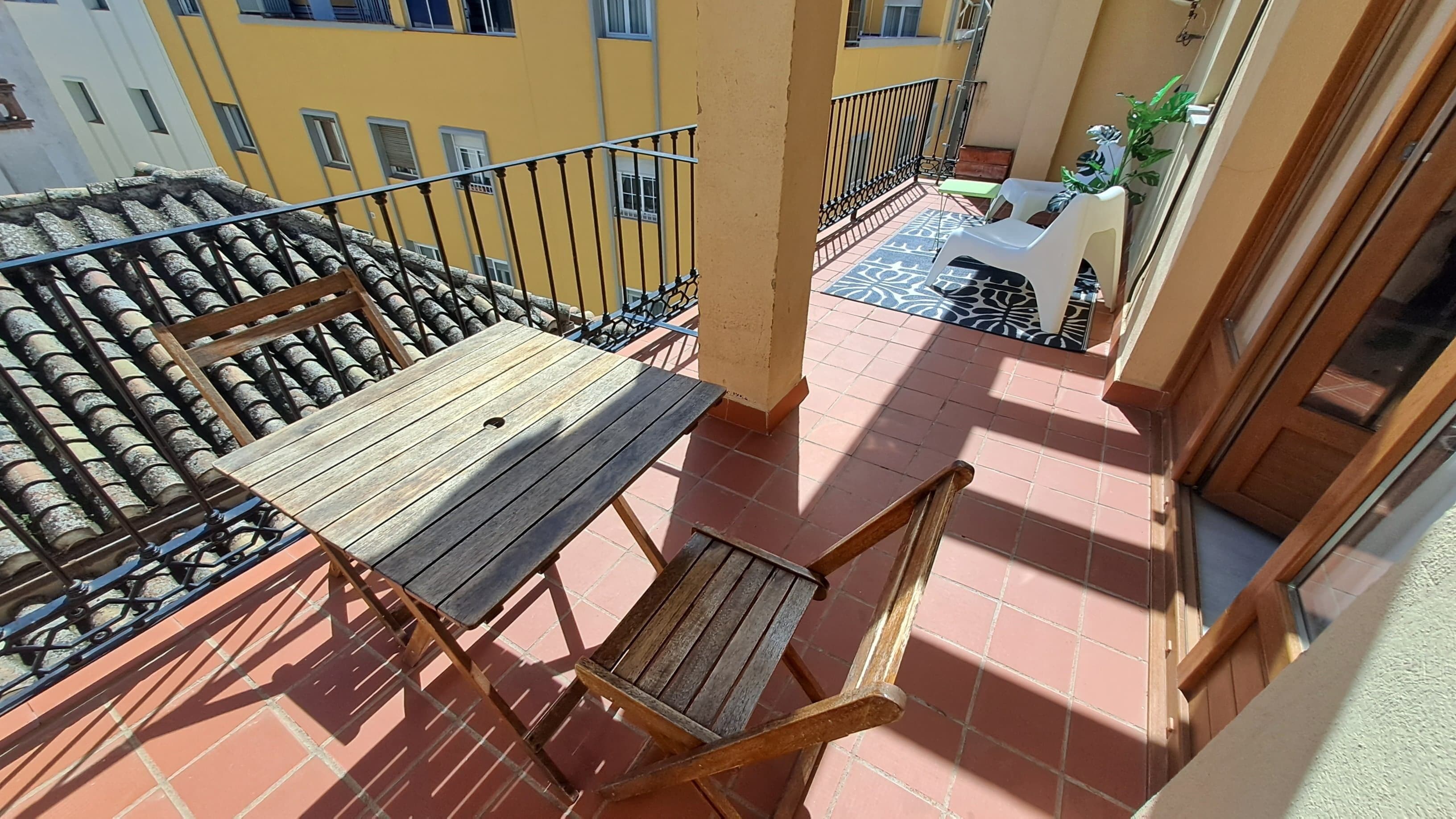 A balcony with a wooden table and a chair, a tiled floor, and a view of a yellow building.