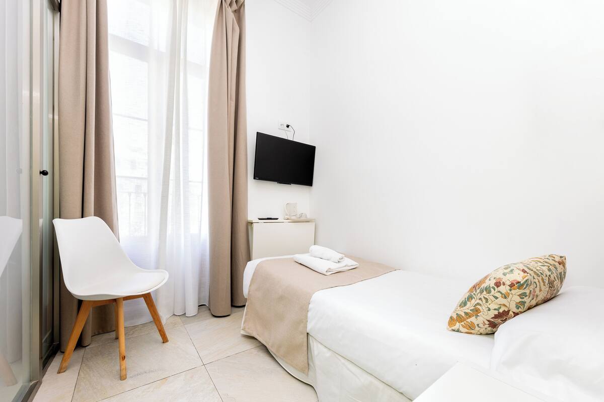 A hotel room with a bed, a chair, a TV, and a window with curtains.