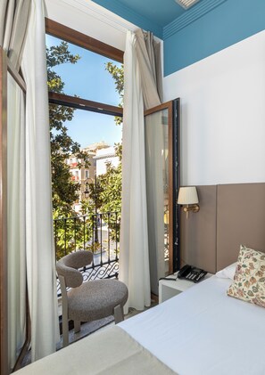 Une chambre d’hôtel avec un lit, une chaise, une table de chevet avec une lampe, et une vue sur la rue et les bâtiments environnants.