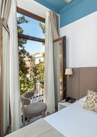 Una habitación de hotel con una cama, una silla, una mesita de noche con una lámpara y vistas a la calle y edificios.