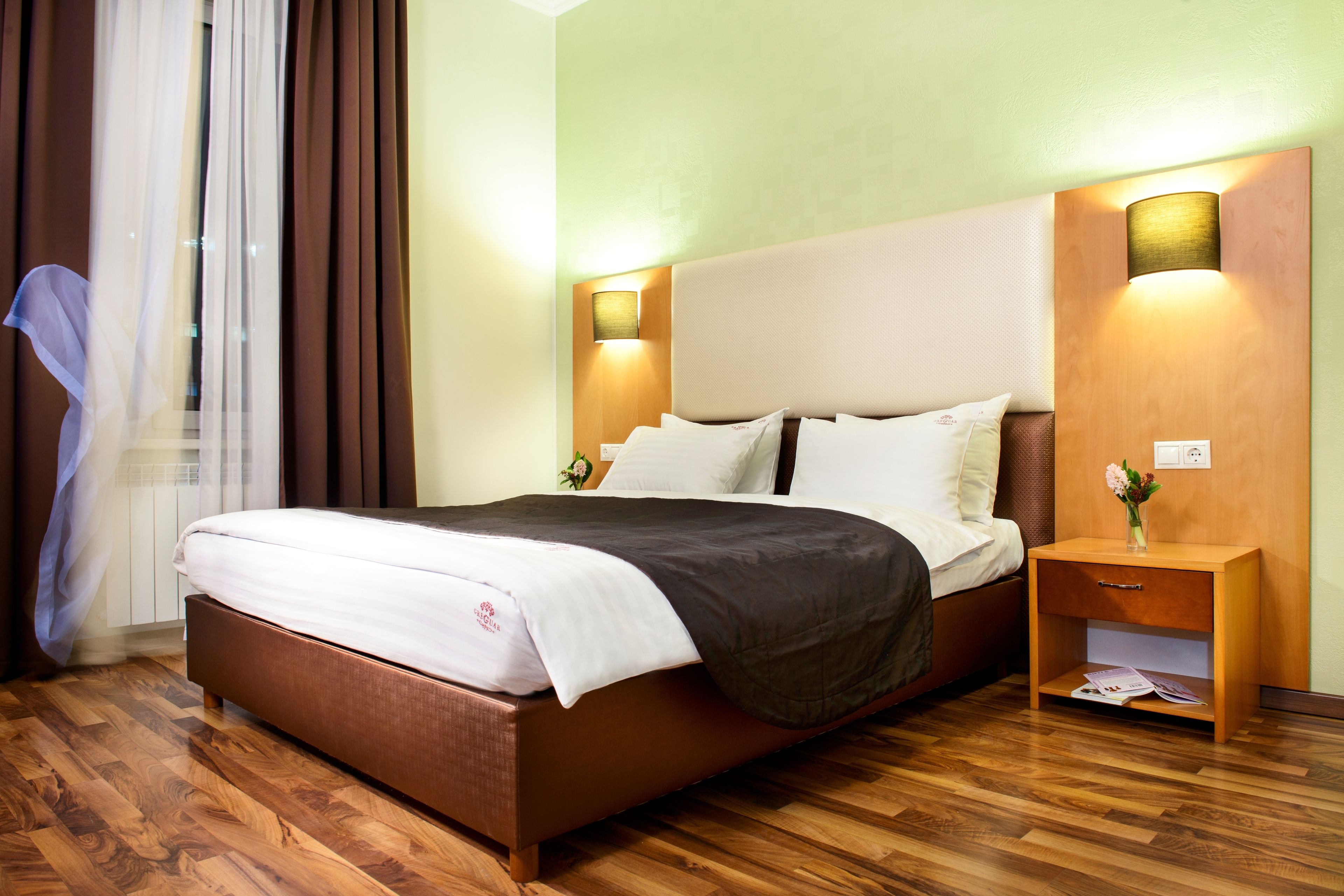 A hotel room with a wooden floor, a bed with white and dark bedding, two bedside lamps, and a nightstand with a vase of flowers.