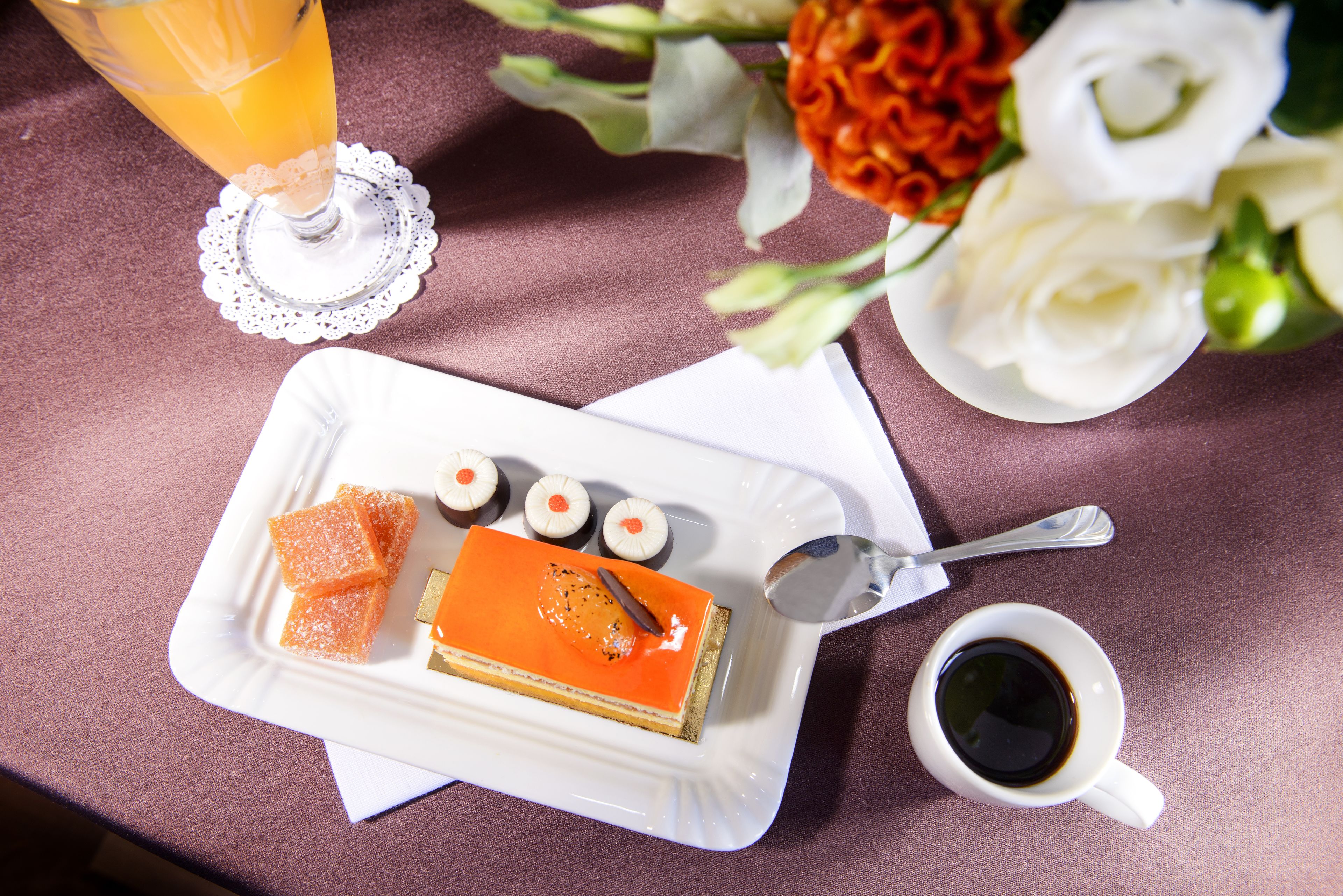 Une assiette à dessert avec une part de gâteau, des cubes d’orange et des bonbons ronds, une tasse de café et un verre de jus d’orange, le tout posé sur une table.