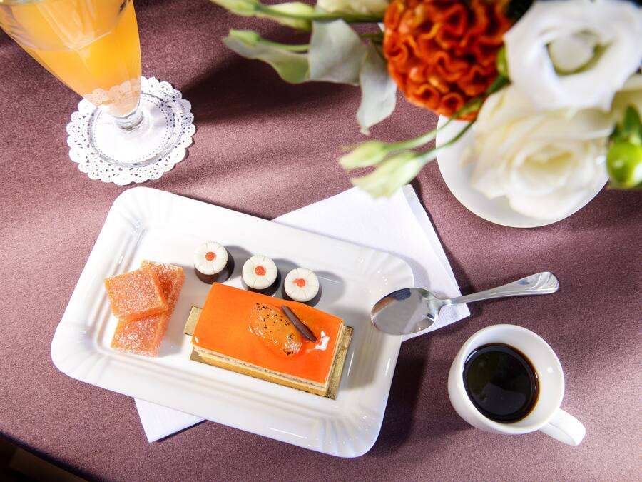 Plat de postre amb una porció de pastís, cubs d'aranja i caramels rodons, una tassa de cafè i un got de suc d'aranja damunt d'una taula.