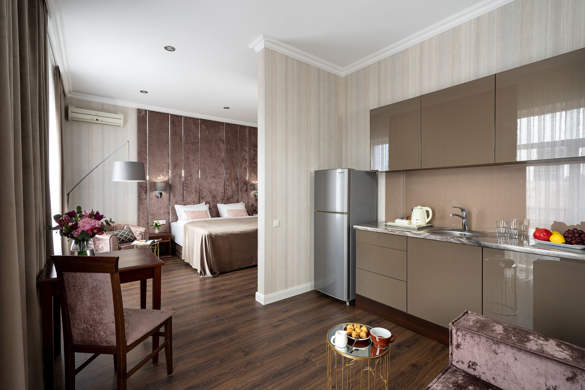A modern hotel room with a wooden floor, a dining area with a table and chairs, a kitchenette with a refrigerator and sink, and a bed with a headboard.