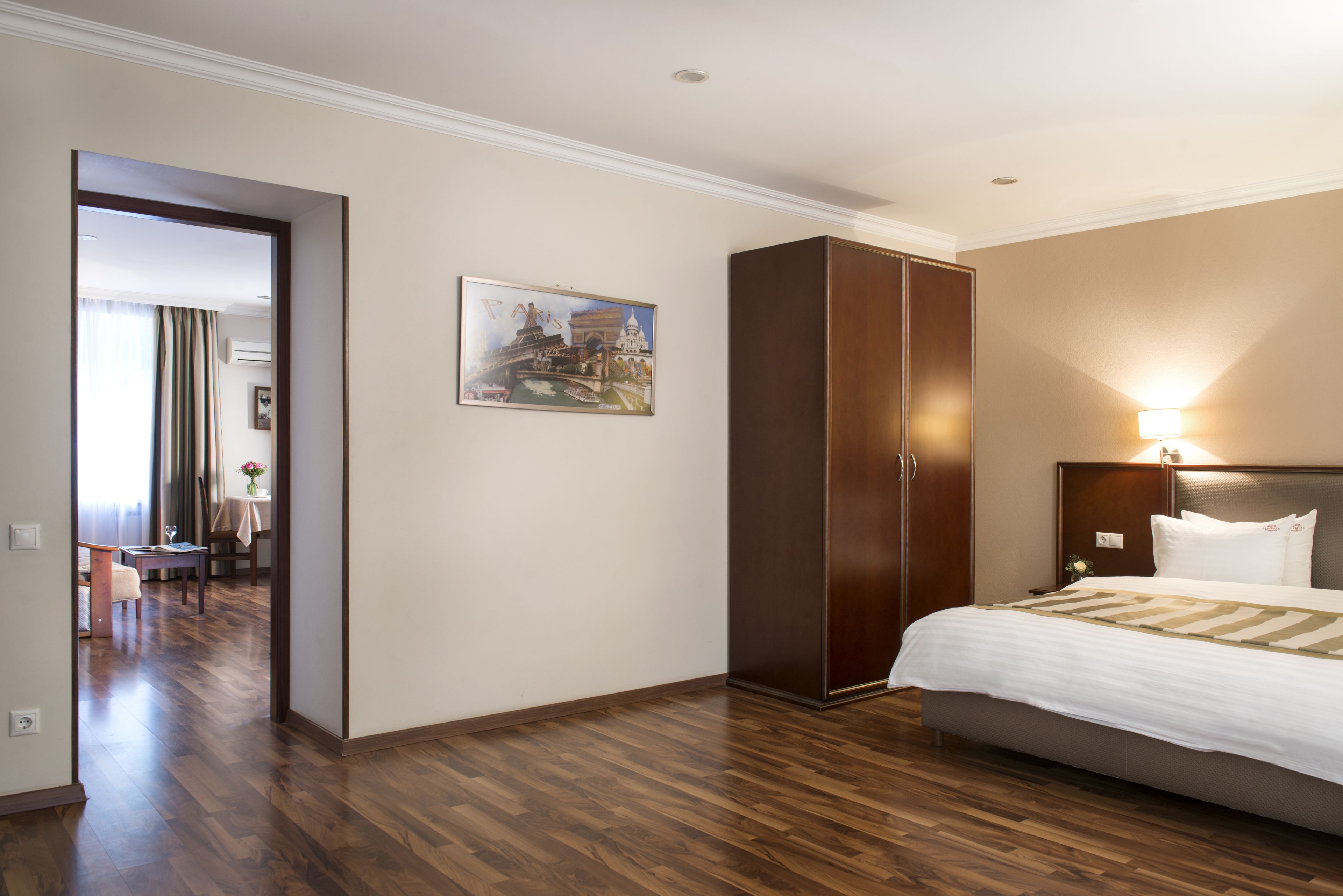 A hotel room with a bed, a wardrobe, a painting, and a dining area in the background.