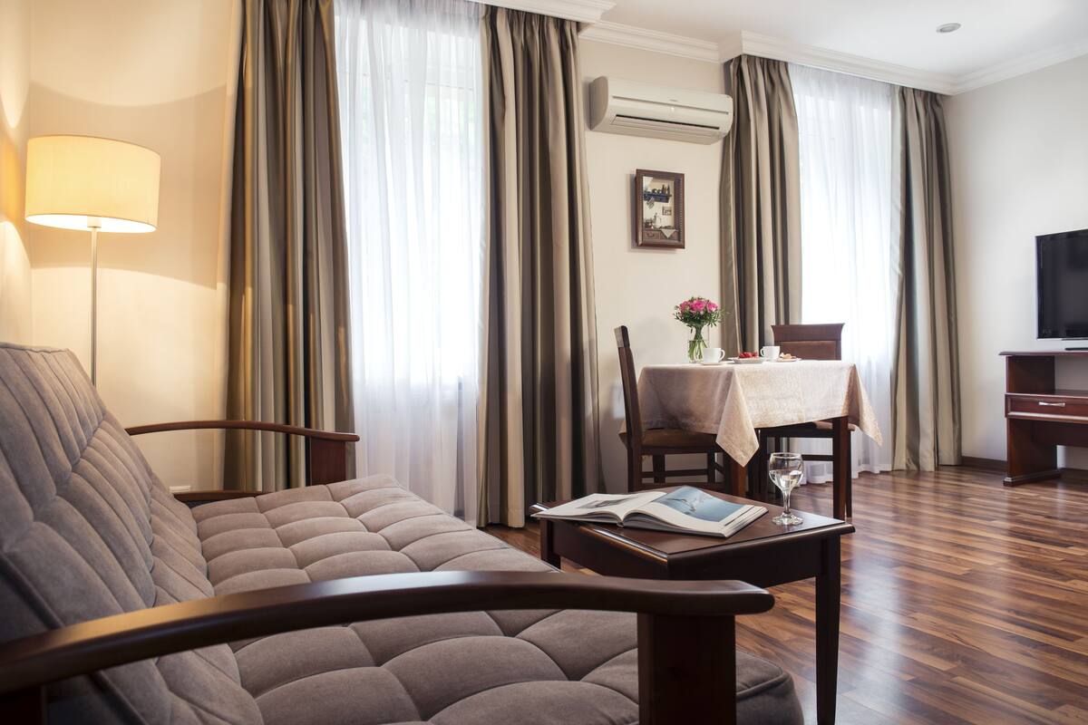 A hotel room with a sofa, a dining table with a vase of flowers, a chair, and a television.