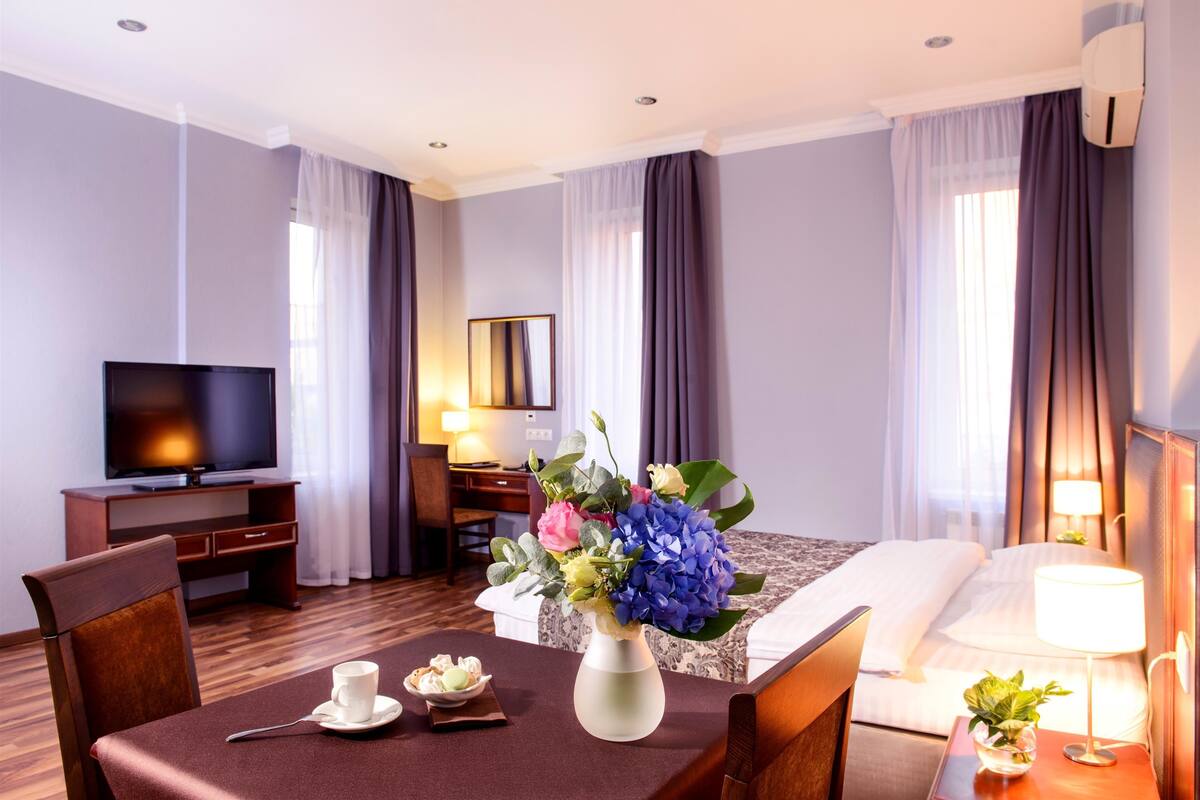 A hotel room with a bed, a desk with a TV, a dining table with a vase of flowers, and a lamp on the bedside table.