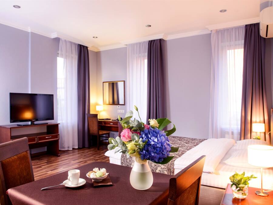 A hotel room with a bed, a desk with a TV, a dining table with a vase of flowers, and a lamp on the bedside table.