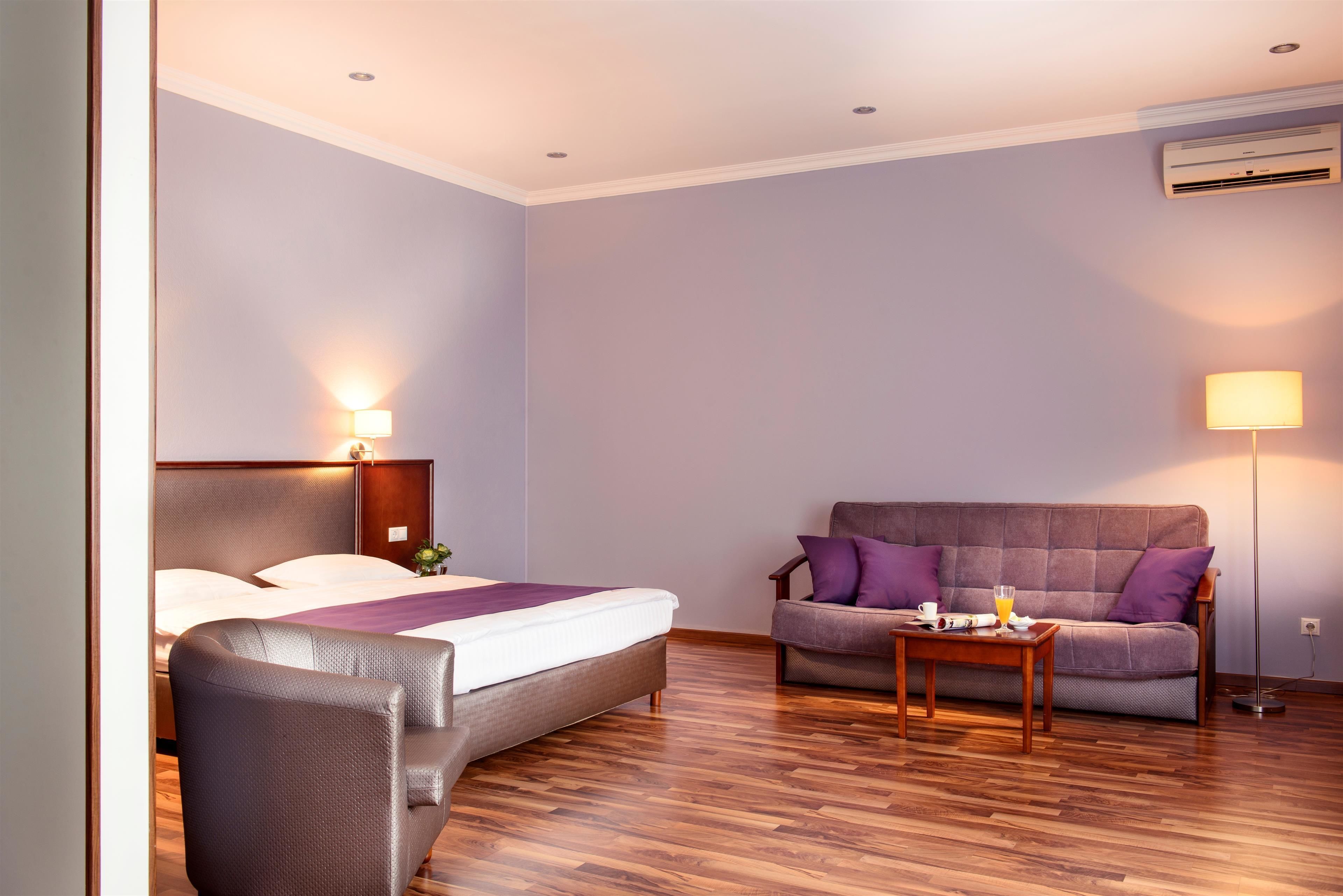 A hotel room with a bed, a sofa, a small table with a cup and a glass, and an air conditioning unit on the wall.