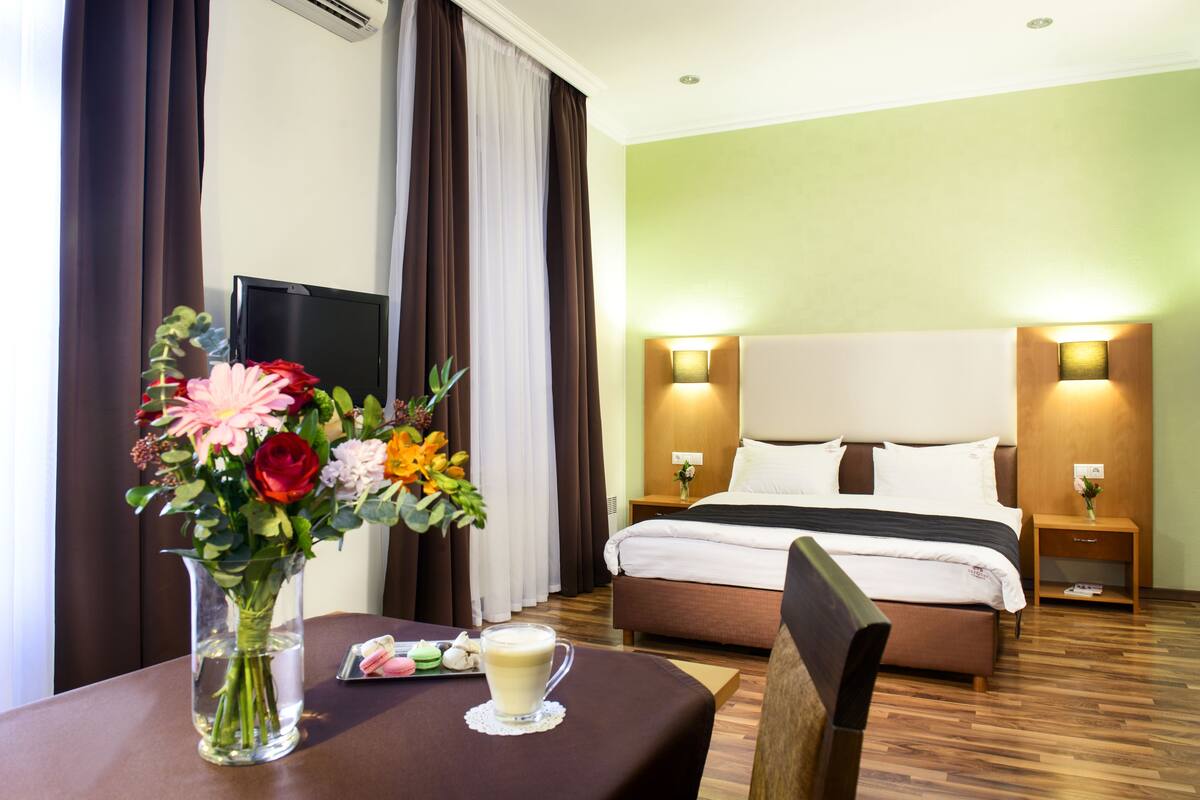 A hotel room with a bed, bedside tables, a TV, a vase with flowers, and a table with a cup and pastries.