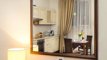 A hotel room with a desk, a lamp, a phone, and a mirror reflecting a dining area with a table, chairs, and a window with curtains.