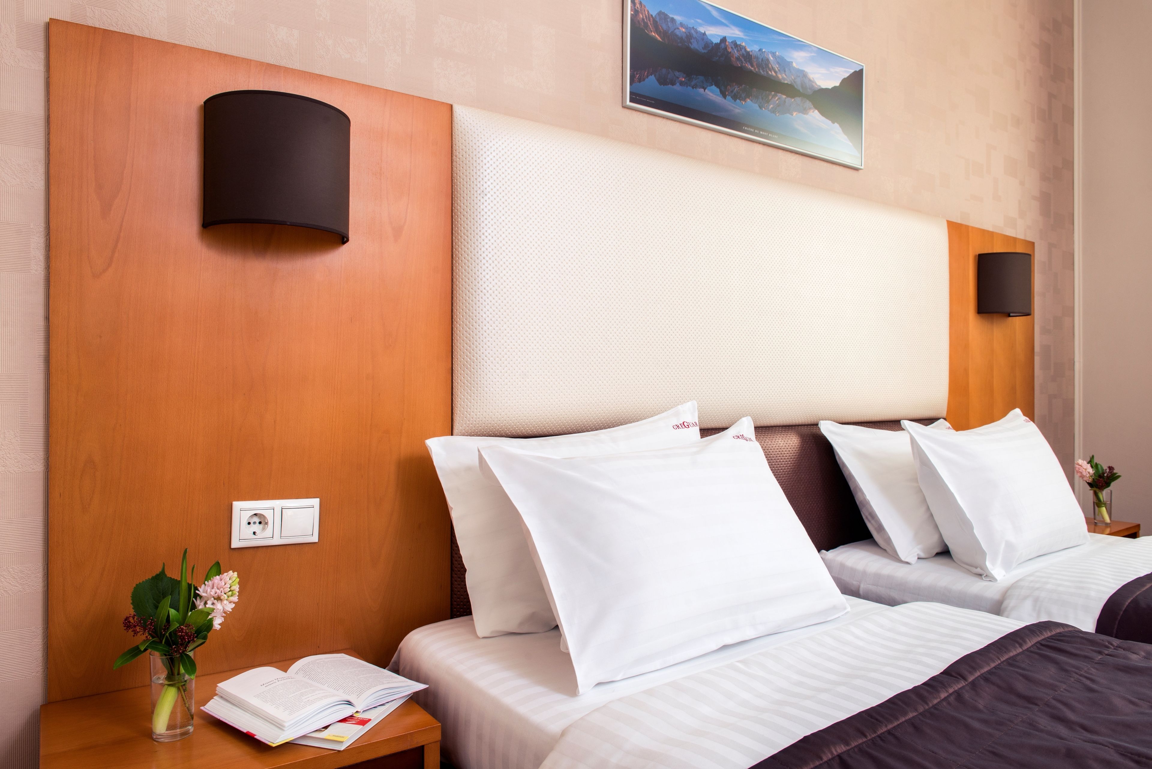 Habitación de hotel con cabecera de madera, dos camas blancas con almohadas a juego, una mesita de noche con un libro y un jarrón de flores, y un televisor montado en la pared.