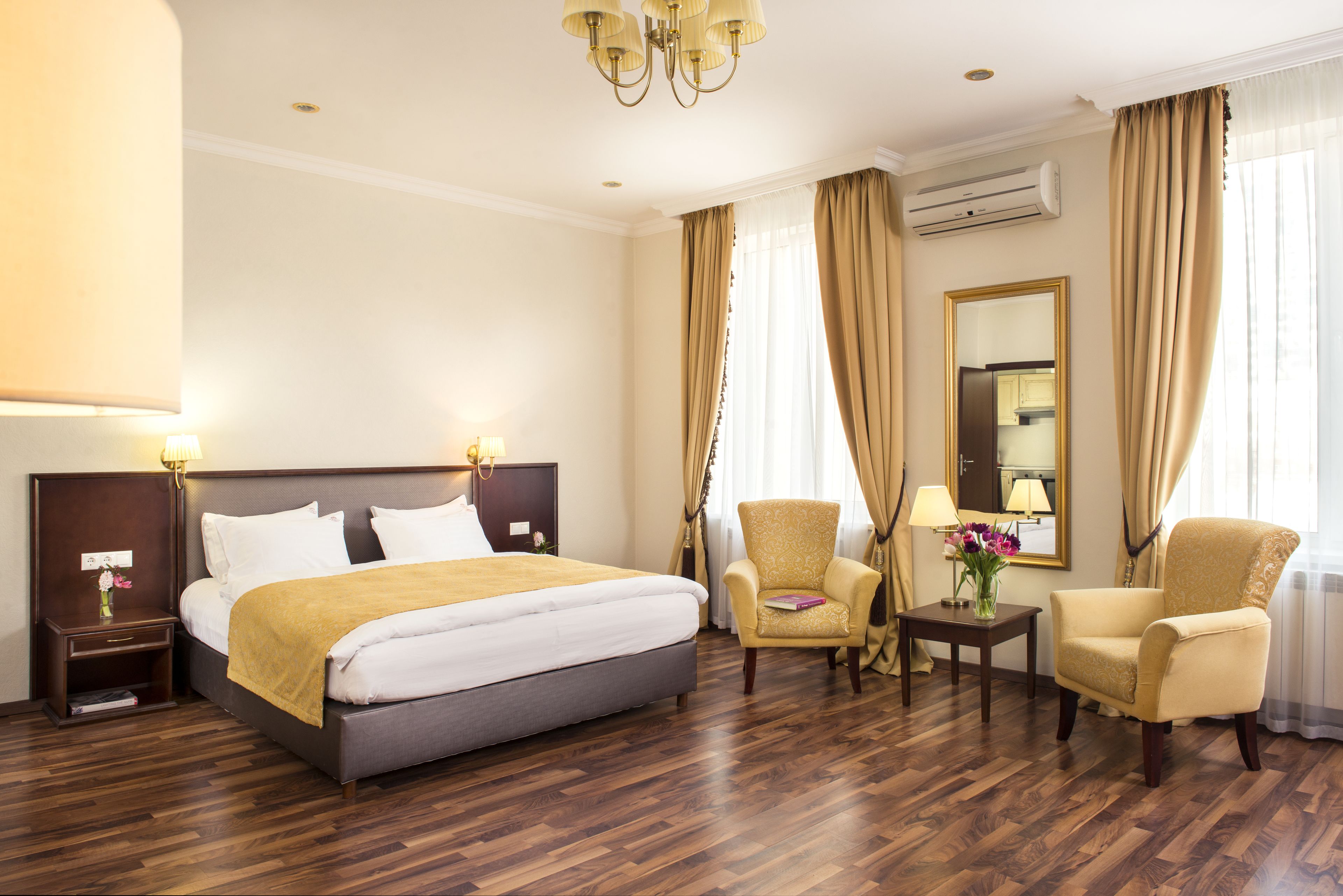 A hotel room with a large bed, two armchairs, a small table with a vase of flowers, and a mirror on the wall.