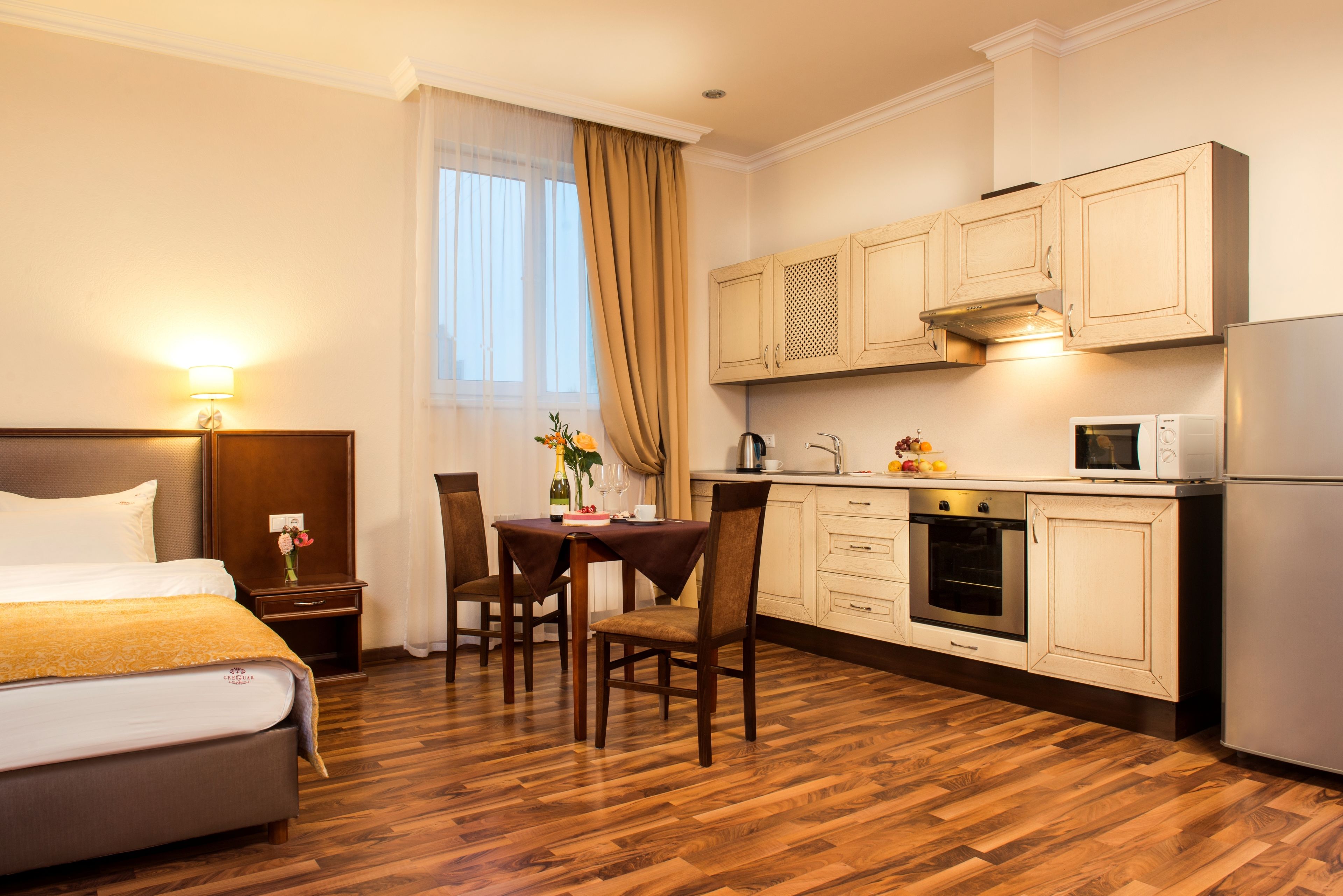 A hotel room with a bed, a dining table with chairs, a kitchen area with cabinets and appliances, and a window with curtains.