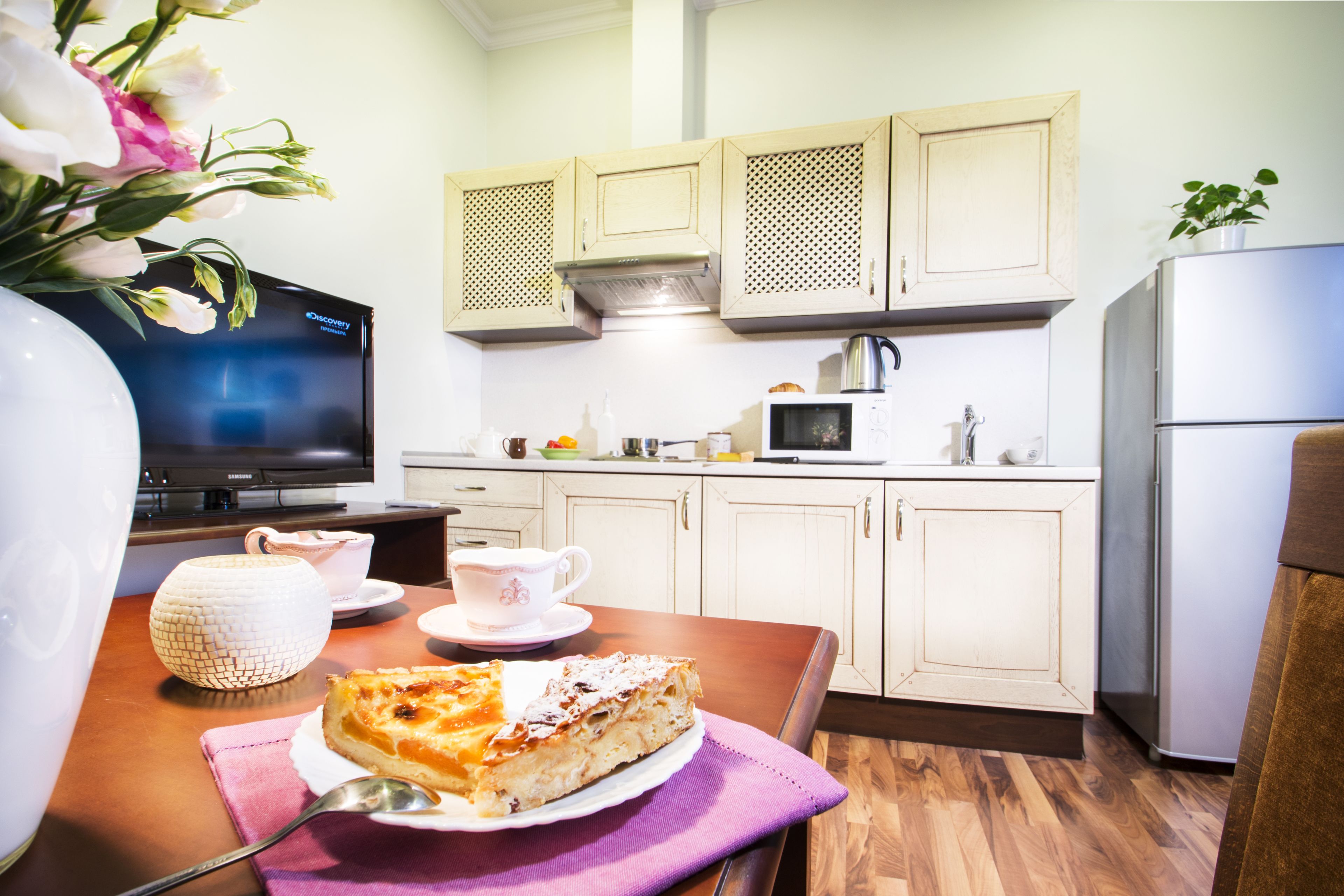 Une cuisine avec des meubles blancs, un four à micro-ondes et un réfrigérateur. Une part de tarte est posée sur une assiette, avec une cuillère sur une serviette de table violette.