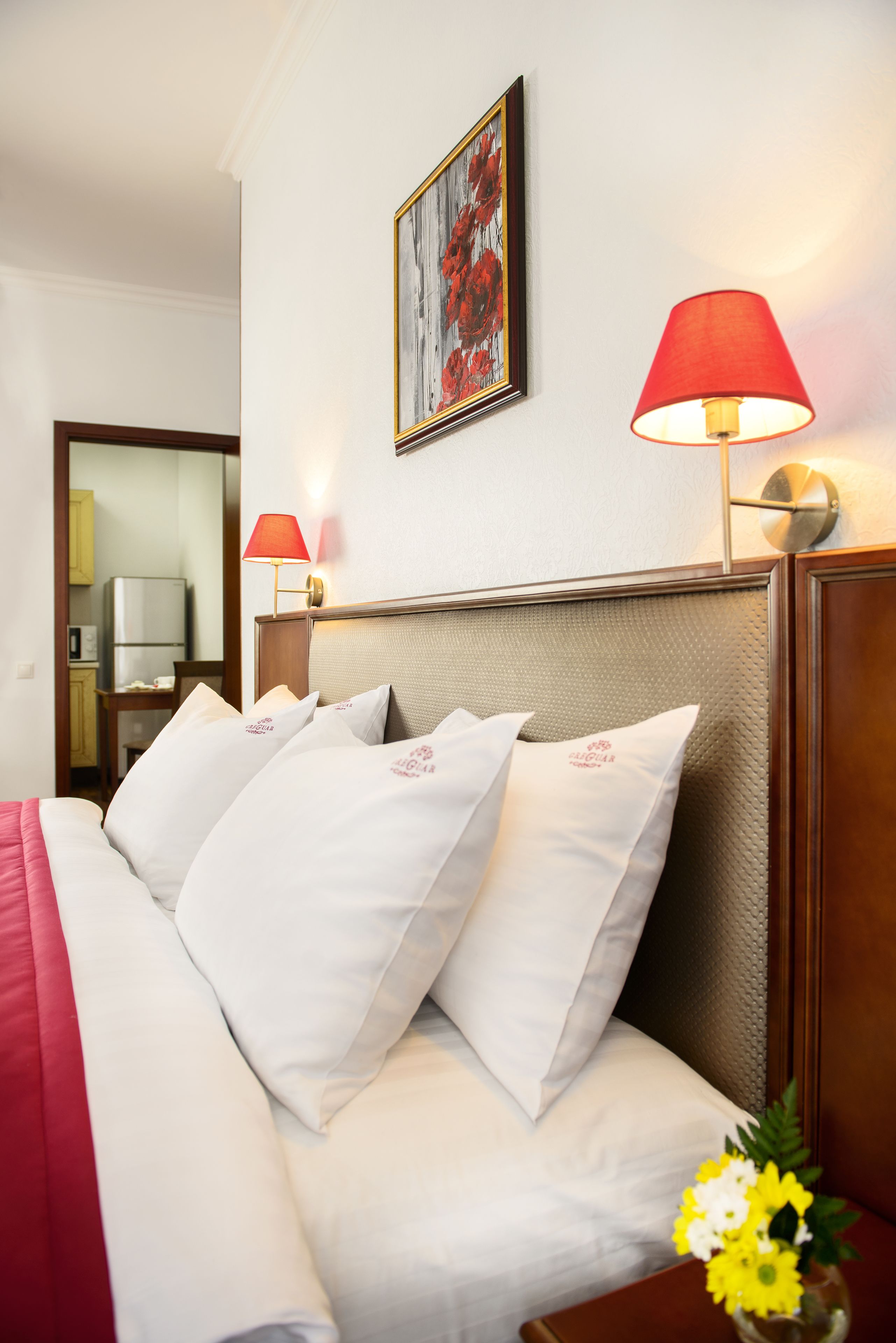 A hotel room with a bed, pillows, a red lamp, a painting on the wall, and a vase with flowers.