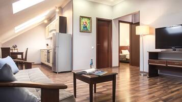 A modern living room with a sofa, a coffee table, and a TV. There is a kitchen area with cabinets and a refrigerator.