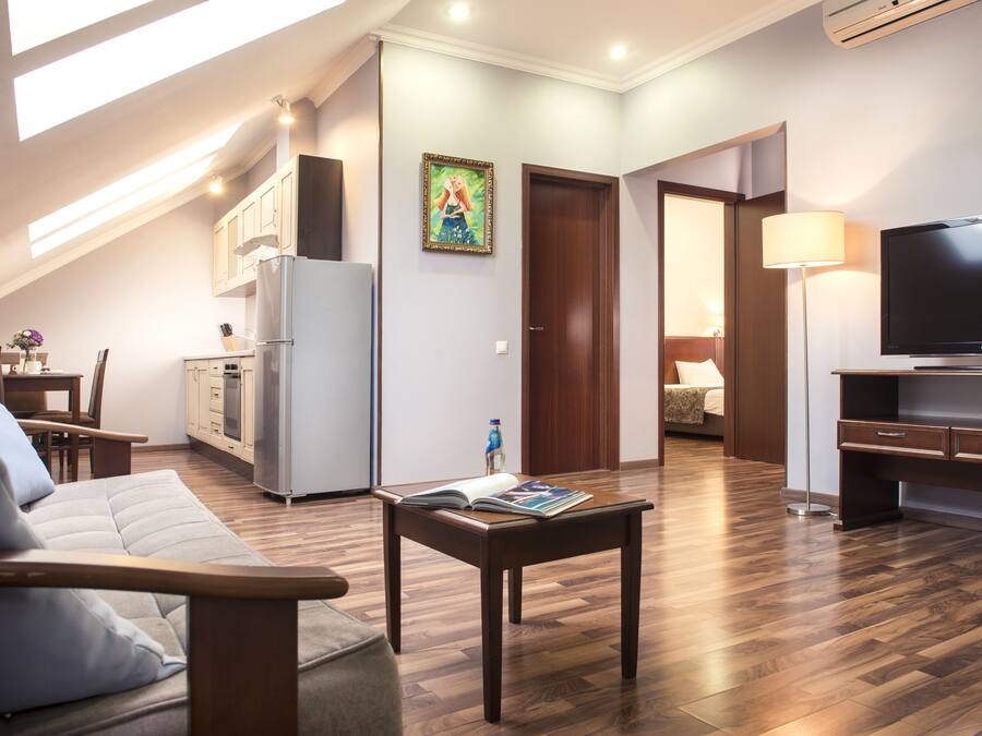 A modern living room with a sofa, a coffee table, and a TV. There is a kitchen area with cabinets and a refrigerator.