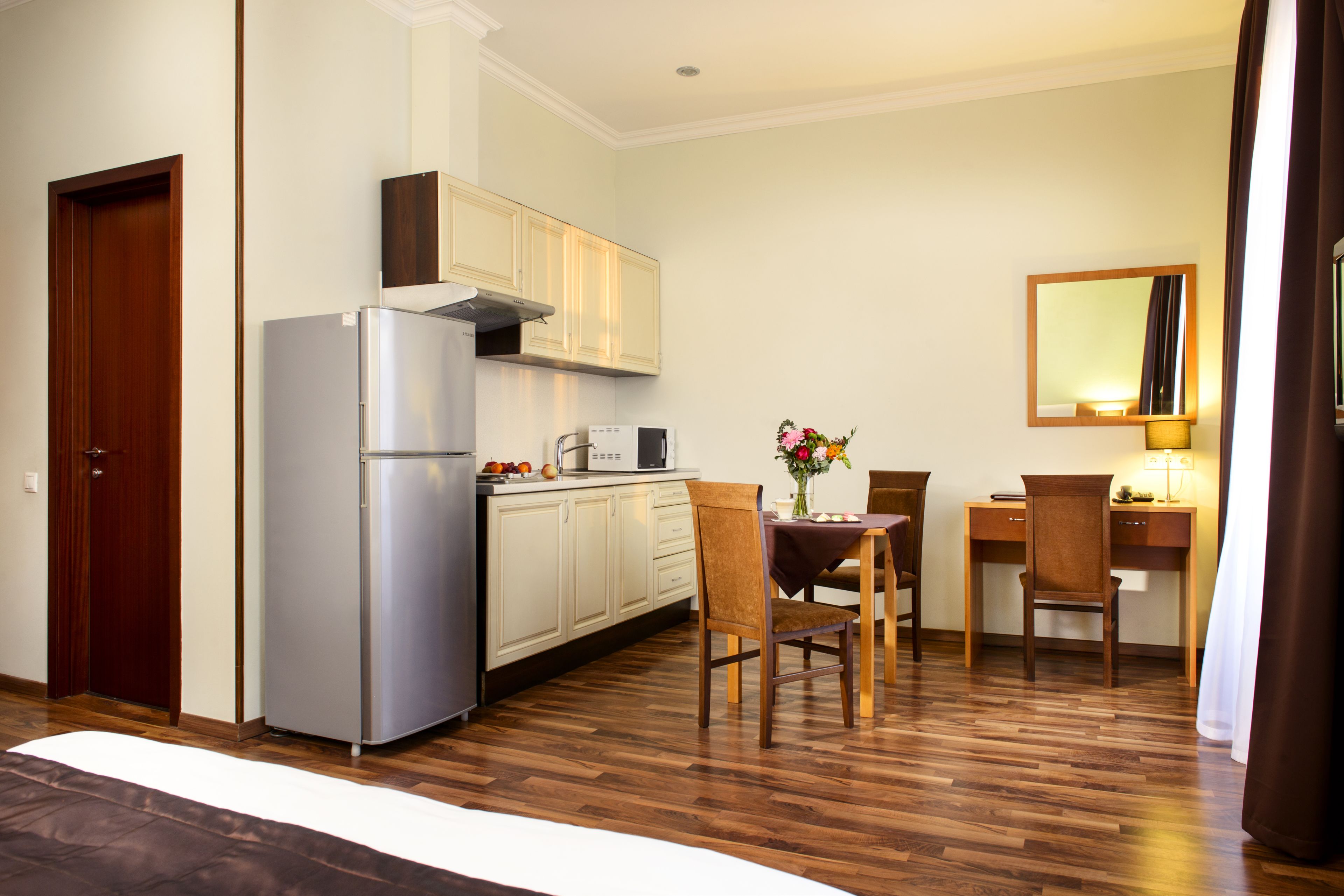 A compact kitchen with a refrigerator, microwave, and wooden cabinets. There is a dining area with a table, chairs, and a vase of flowers.