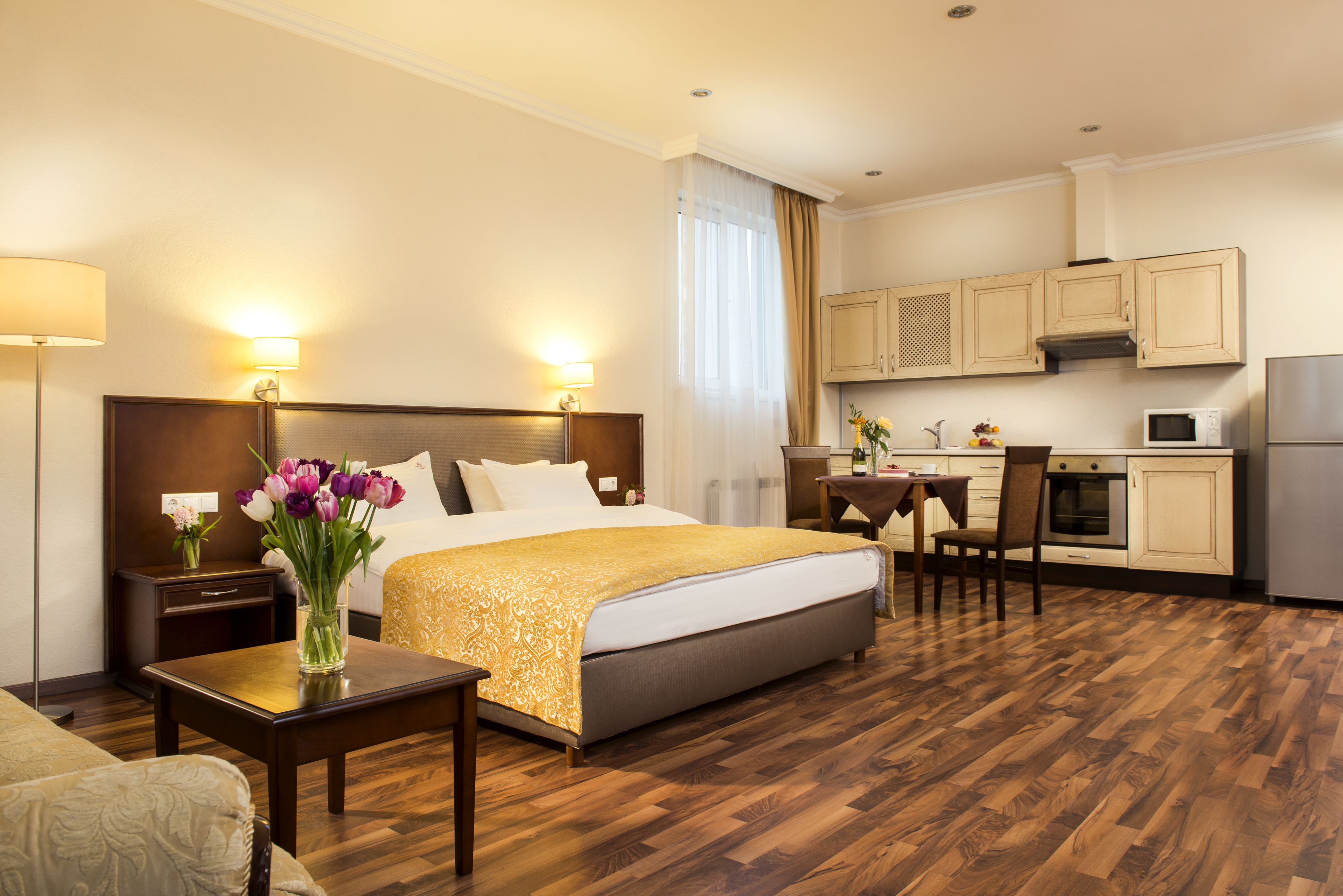 Habitación de hotel con cama, mesitas de noche, una pequeña mesa con un florero, un área de comedor con mesa y sillas, y una cocina con armarios y electrodomésticos.