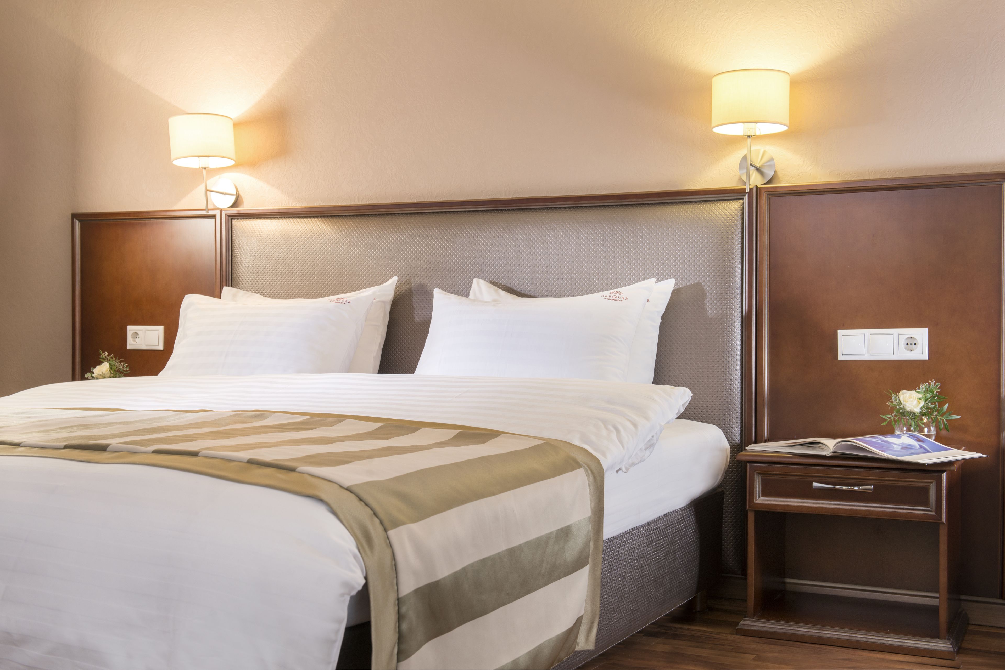 A neatly made bed with white linens and a striped throw blanket, a bedside table with a book and a vase of flowers, and two wall-mounted lamps.