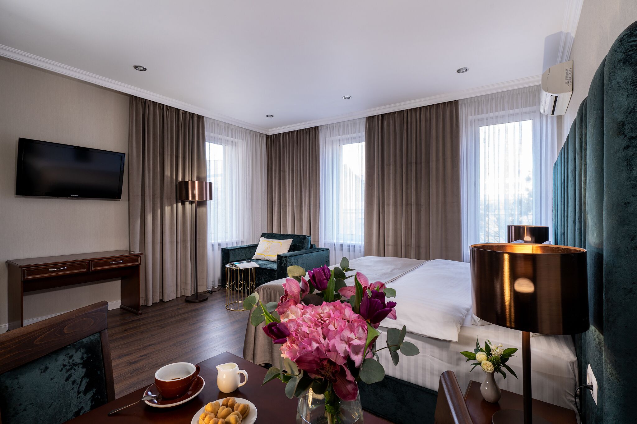 A hotel room with a bed, a desk, a TV, a lamp, and a dining table with a vase of flowers.