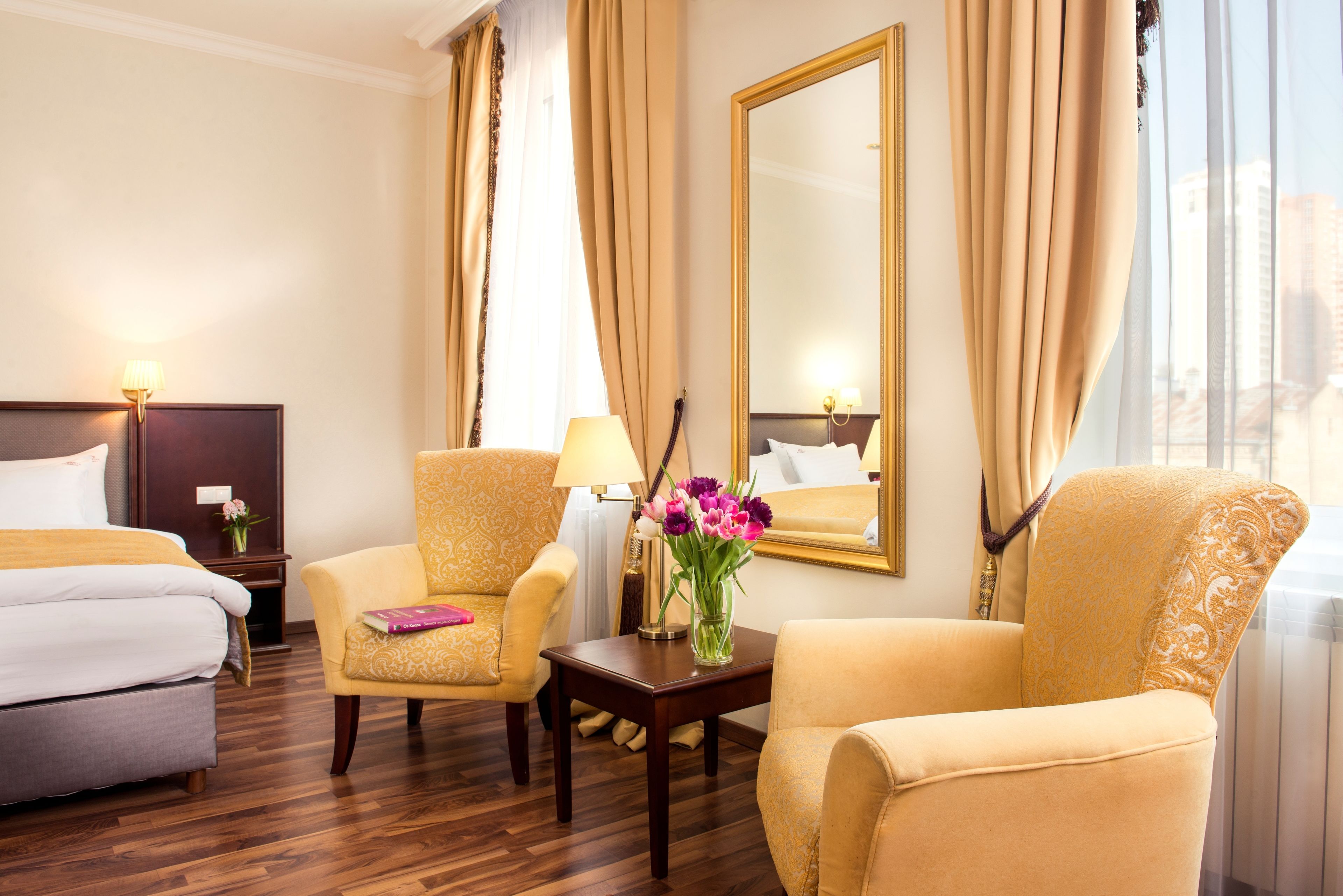 A hotel room with a bed, two armchairs, a small table with a vase of flowers, and a large mirror on the wall.