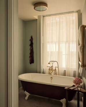 Una bañera clásica tipo pata de león con grifo dorado, una ventana con cortinas de gasa y una mesita de madera con un florero.