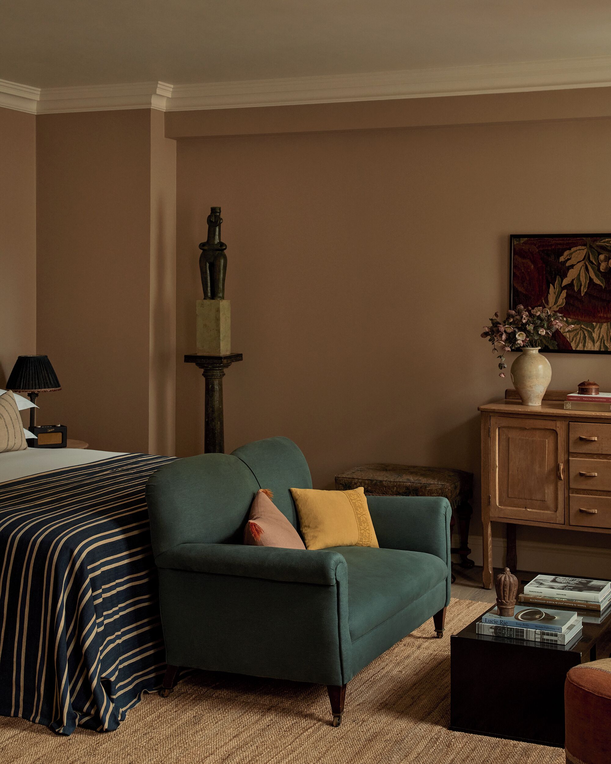 Una habitación con una cama, un sillón verde, un armario de madera y una mesita con libros.