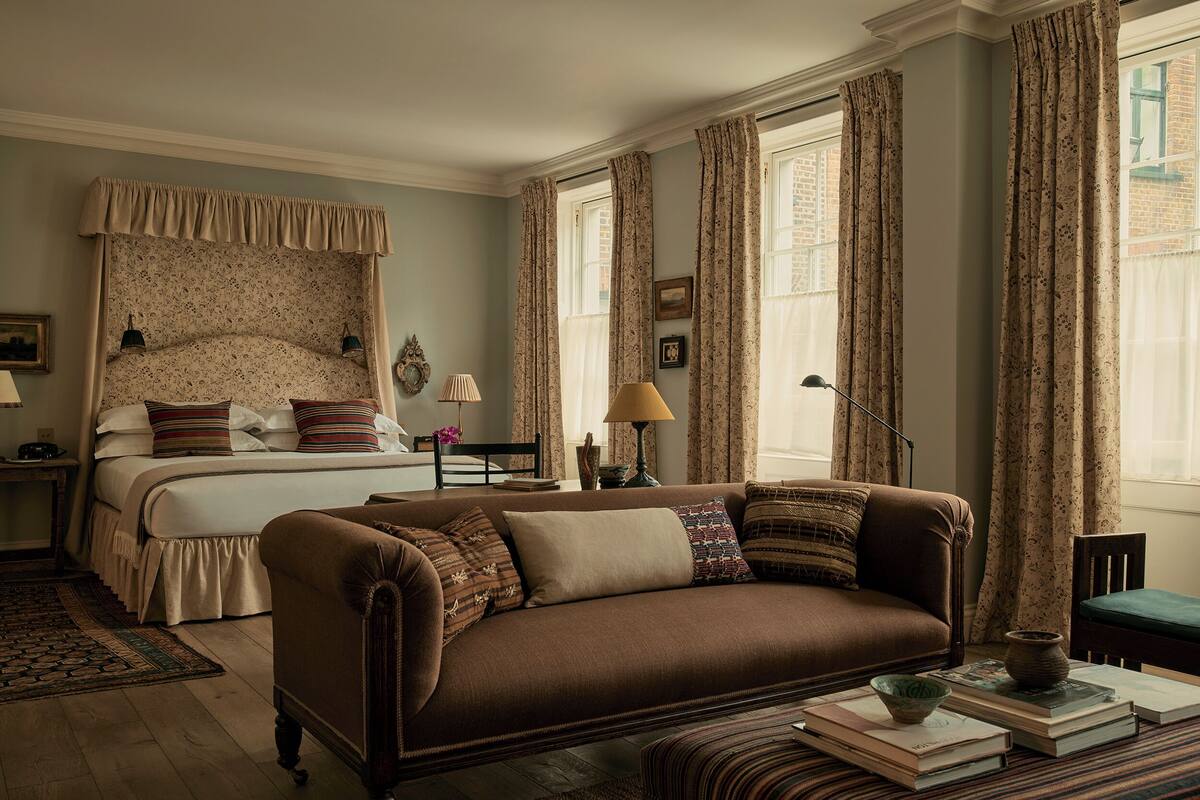 A bedroom with a bed, a brown sofa, a bedside table, a lamp, and a window with curtains.