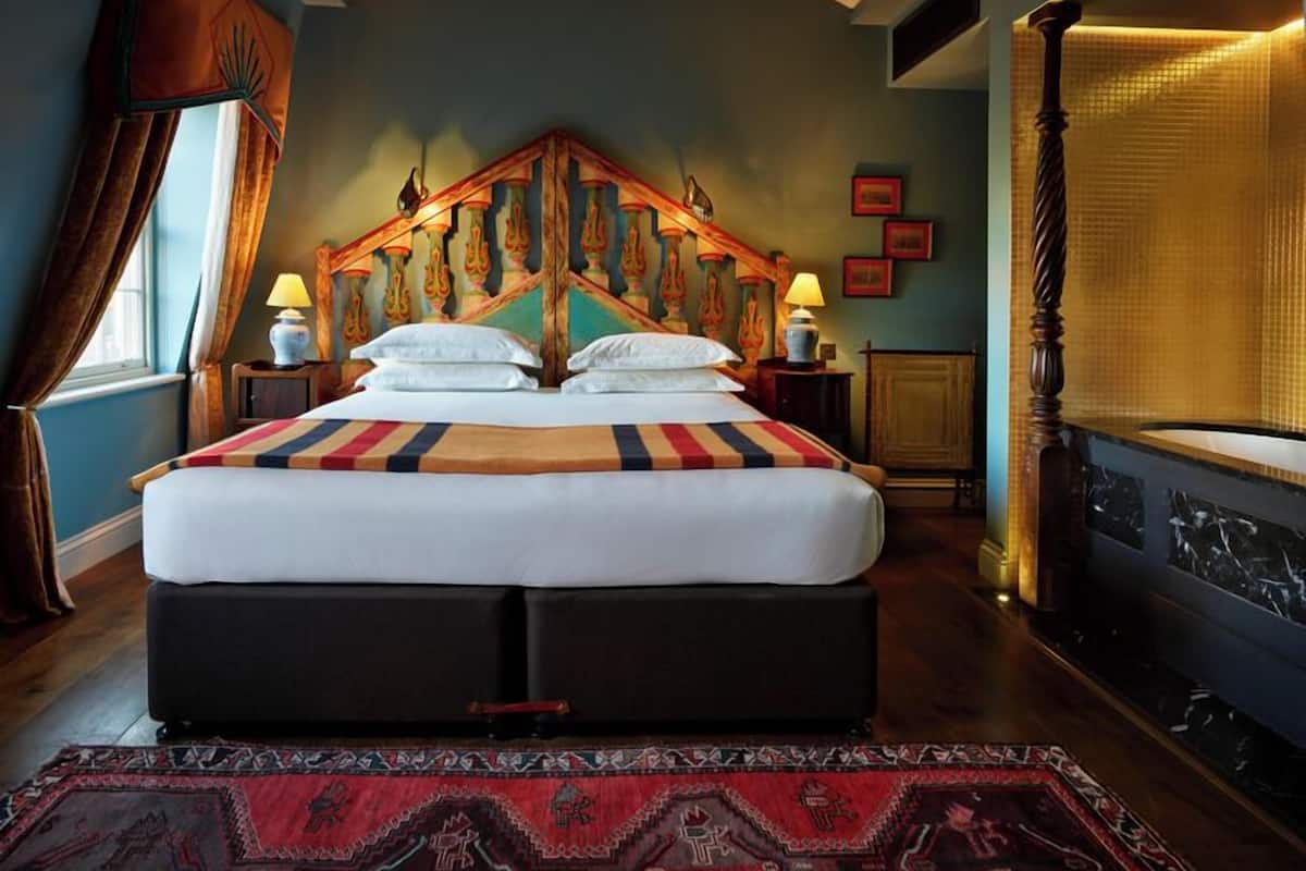 A large bed with a colorful headboard, two bedside lamps, a patterned rug, and a window with curtains.