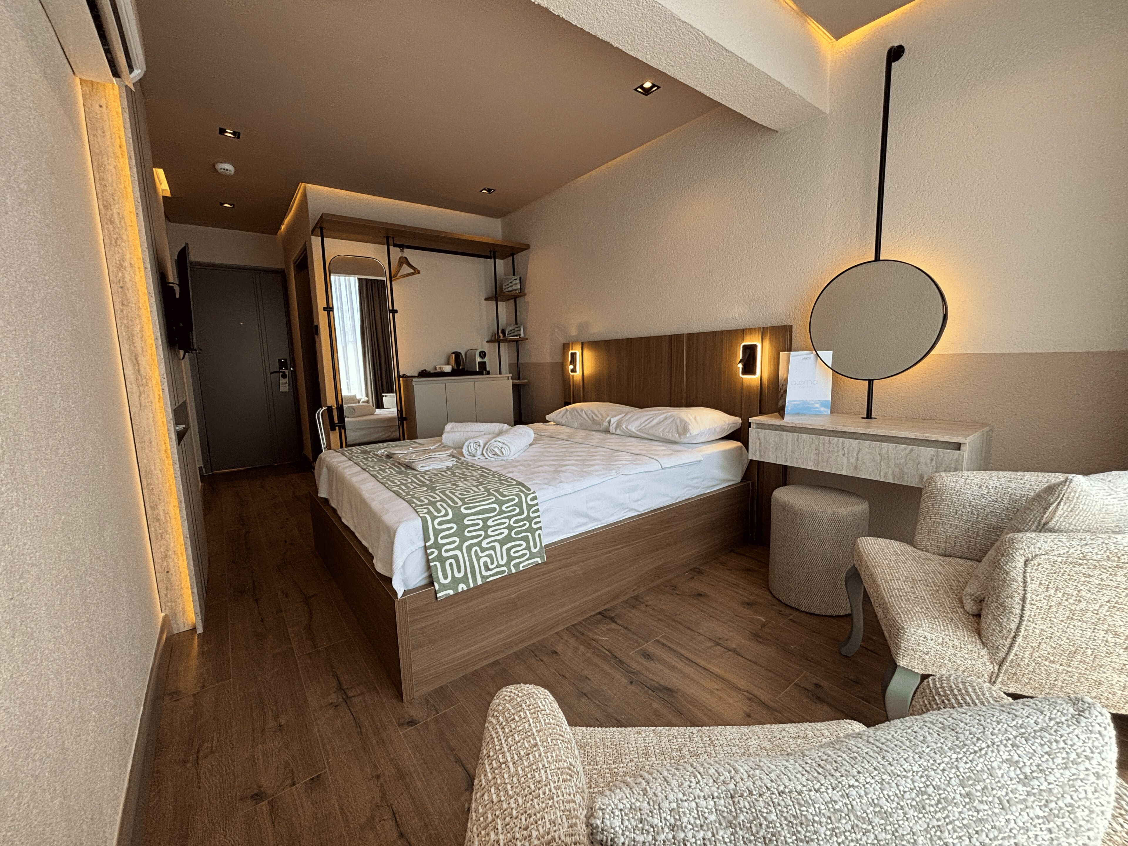 A hotel room with a wooden bed, a round mirror, a small seating area, and a kitchenette.