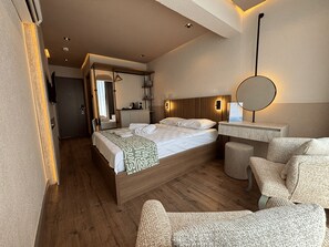 A hotel room with a wooden bed, a round mirror, a small seating area, and a kitchenette. - Aterna Hotel (Dikili)
