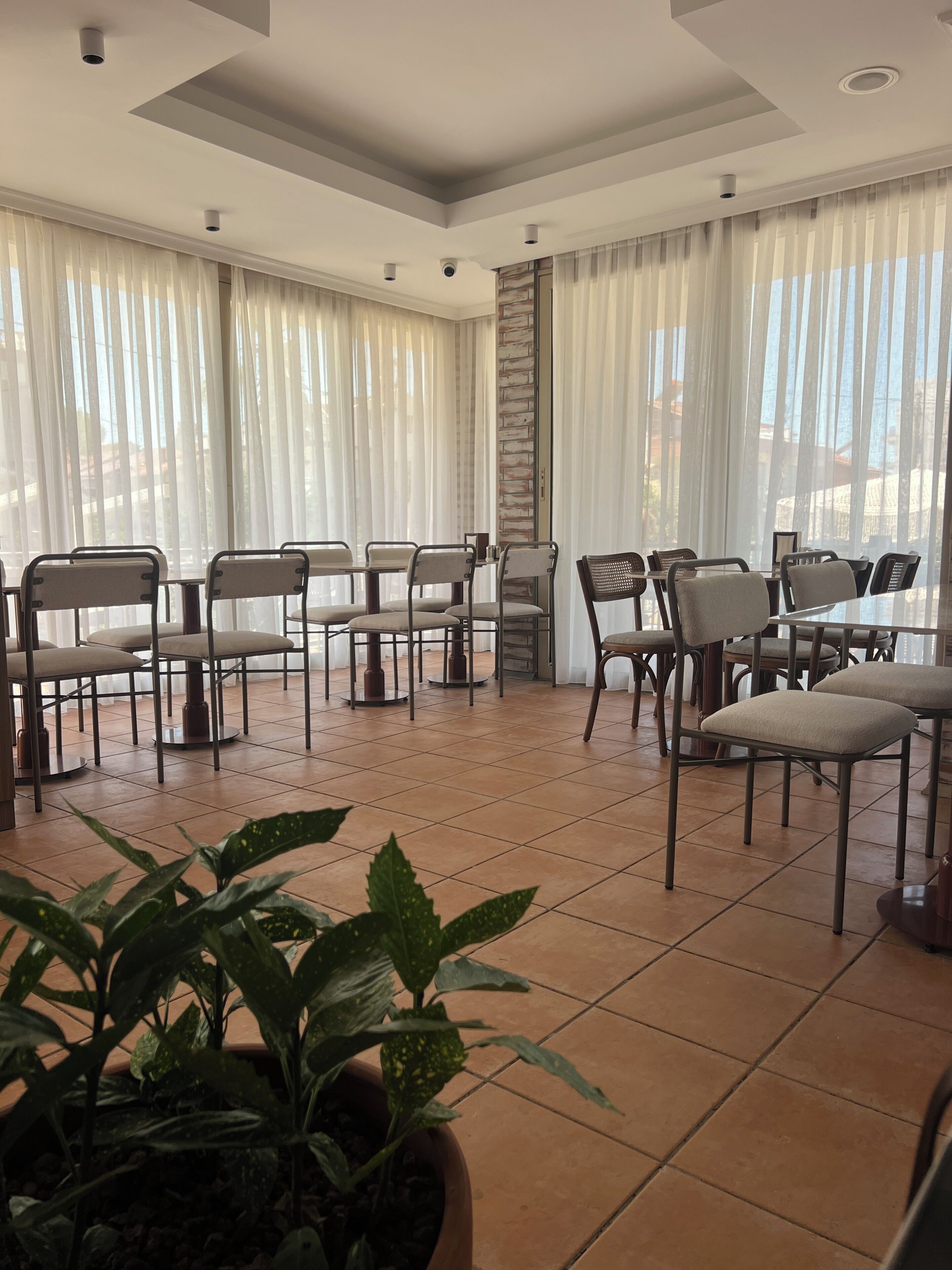 Un comedor con sillas y mesas, ventanas amplias con cortinas y piso de baldosas.